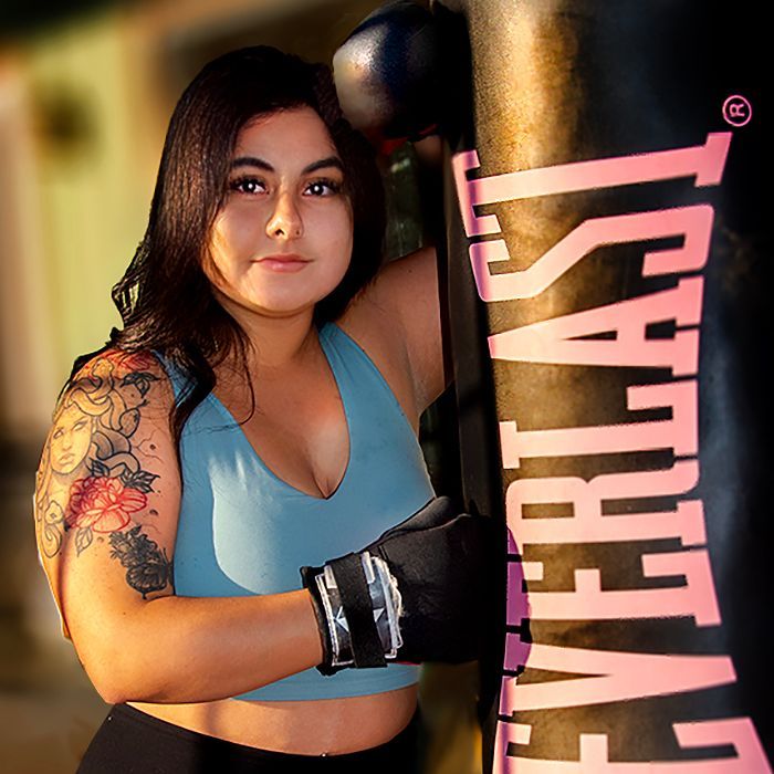 Woman in blue sports bra and boxing gloves leaning on Everlast punching bag.