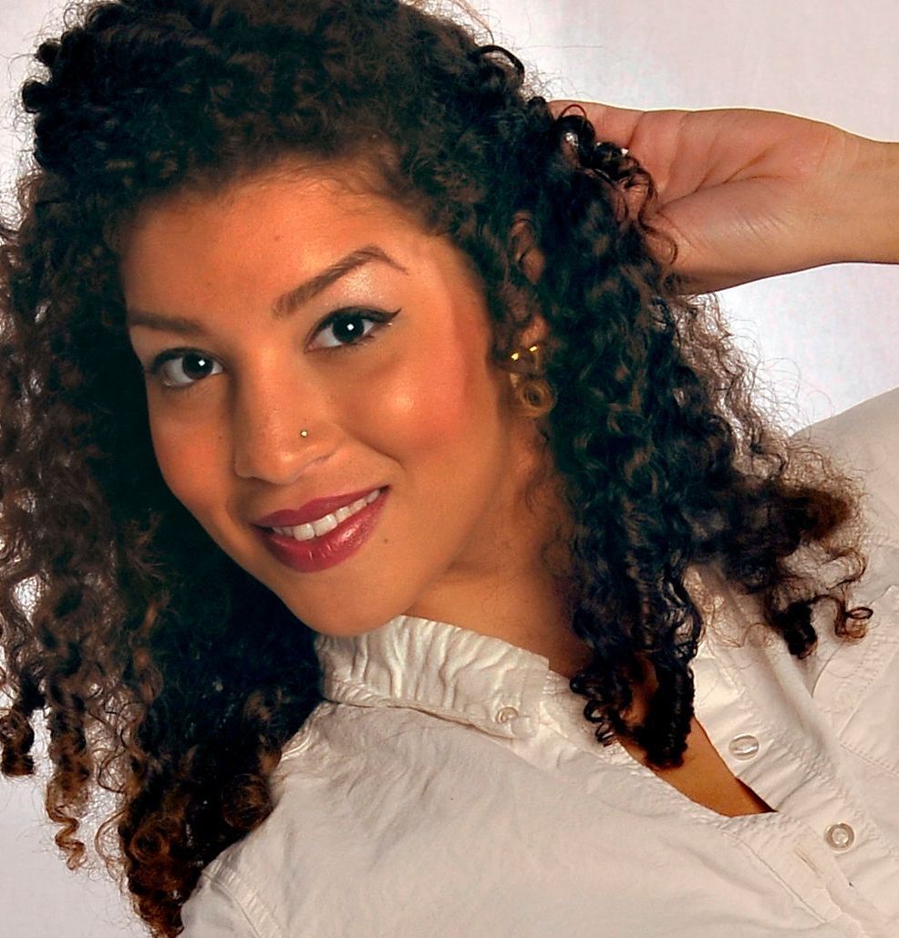 Woman with curly dark hair, smiling, wearing a white shirt, hand in hair.
