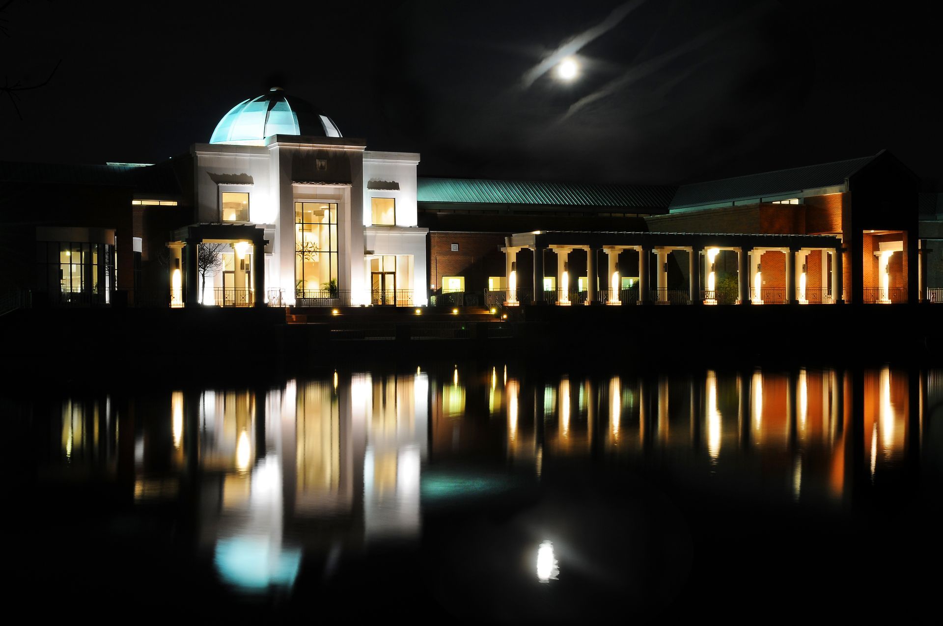 Night view of a building with a domed roof and colonnade, reflected in calm water under a dark sky with the moon.