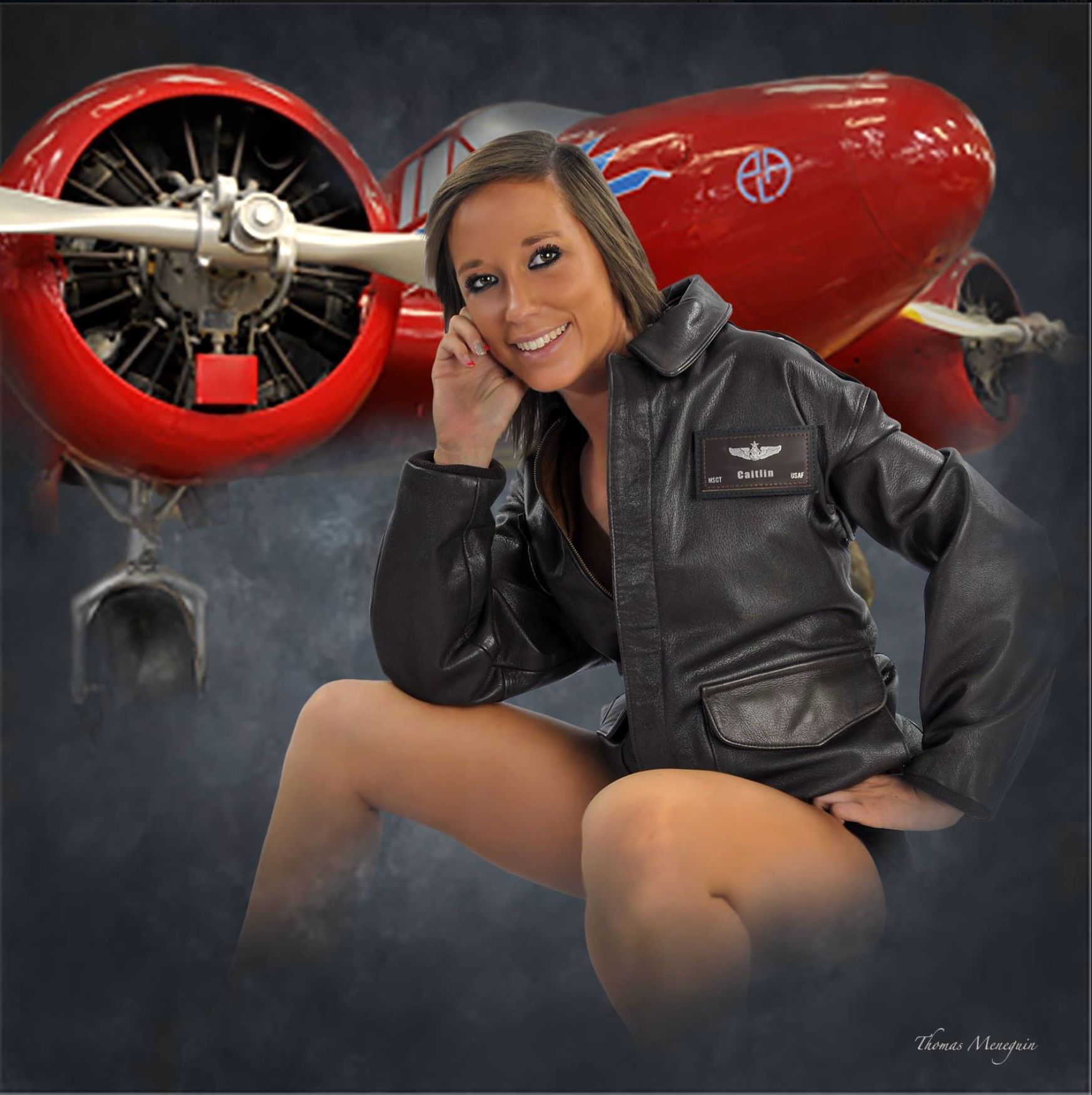Woman in leather aviator jacket, seated near a vintage red airplane, smiling.