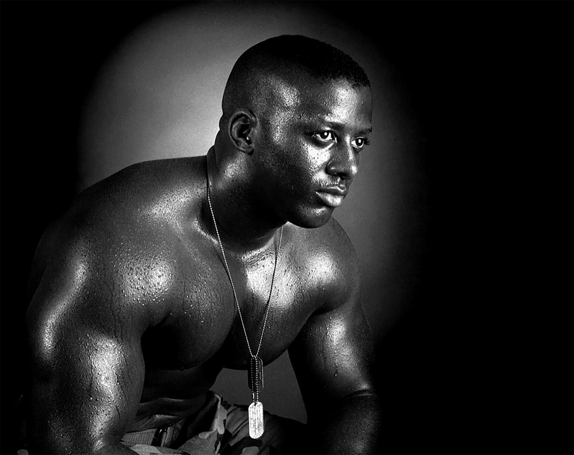 Sweaty, muscular person wearing a dog tag, looking off to the side, seated, under a spotlight.