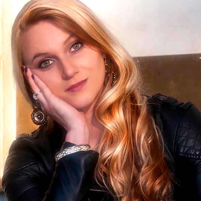Woman with long blonde hair, resting chin on hand, wearing a black leather jacket.