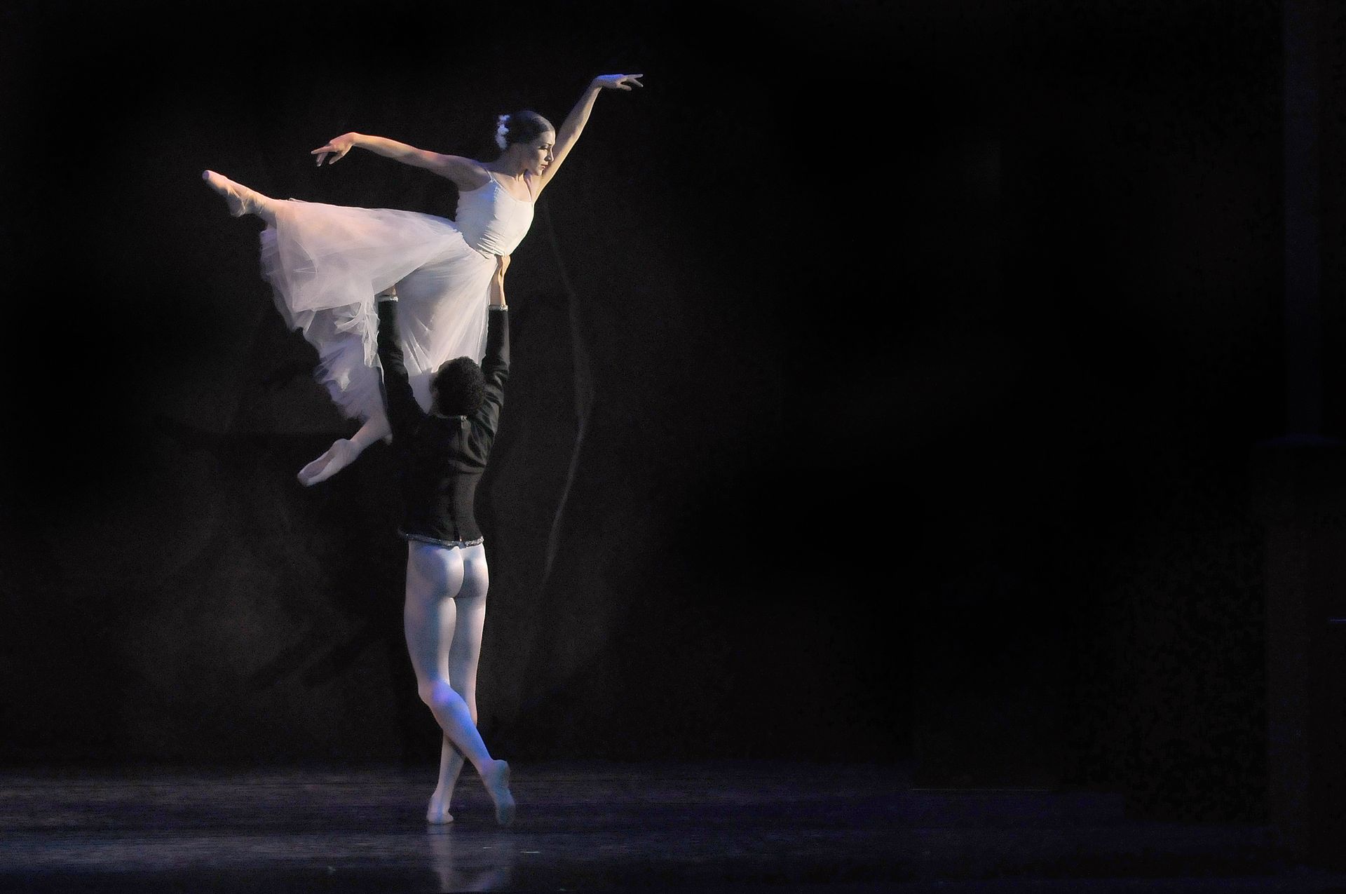Ballet dancers performing a lift on stage. The woman is held aloft, wearing white; man in black.