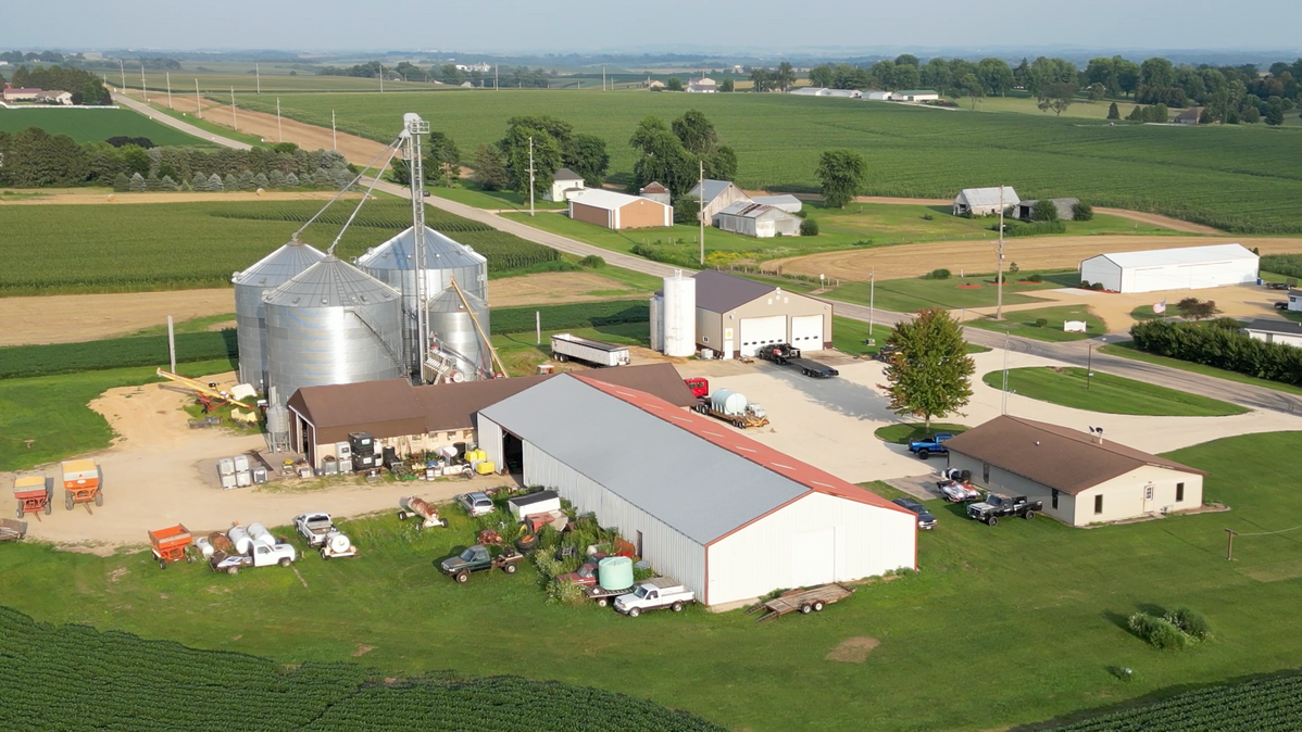 Farm complex with silver grain silos, various buildings, fields, and a road.