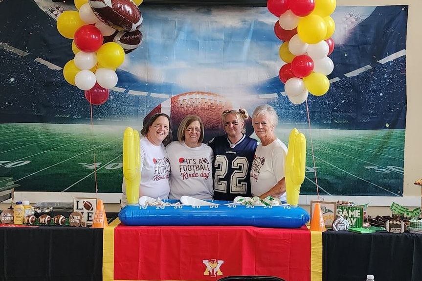 Four people pose at a football-themed party table, decorated with balloons and a backdrop.