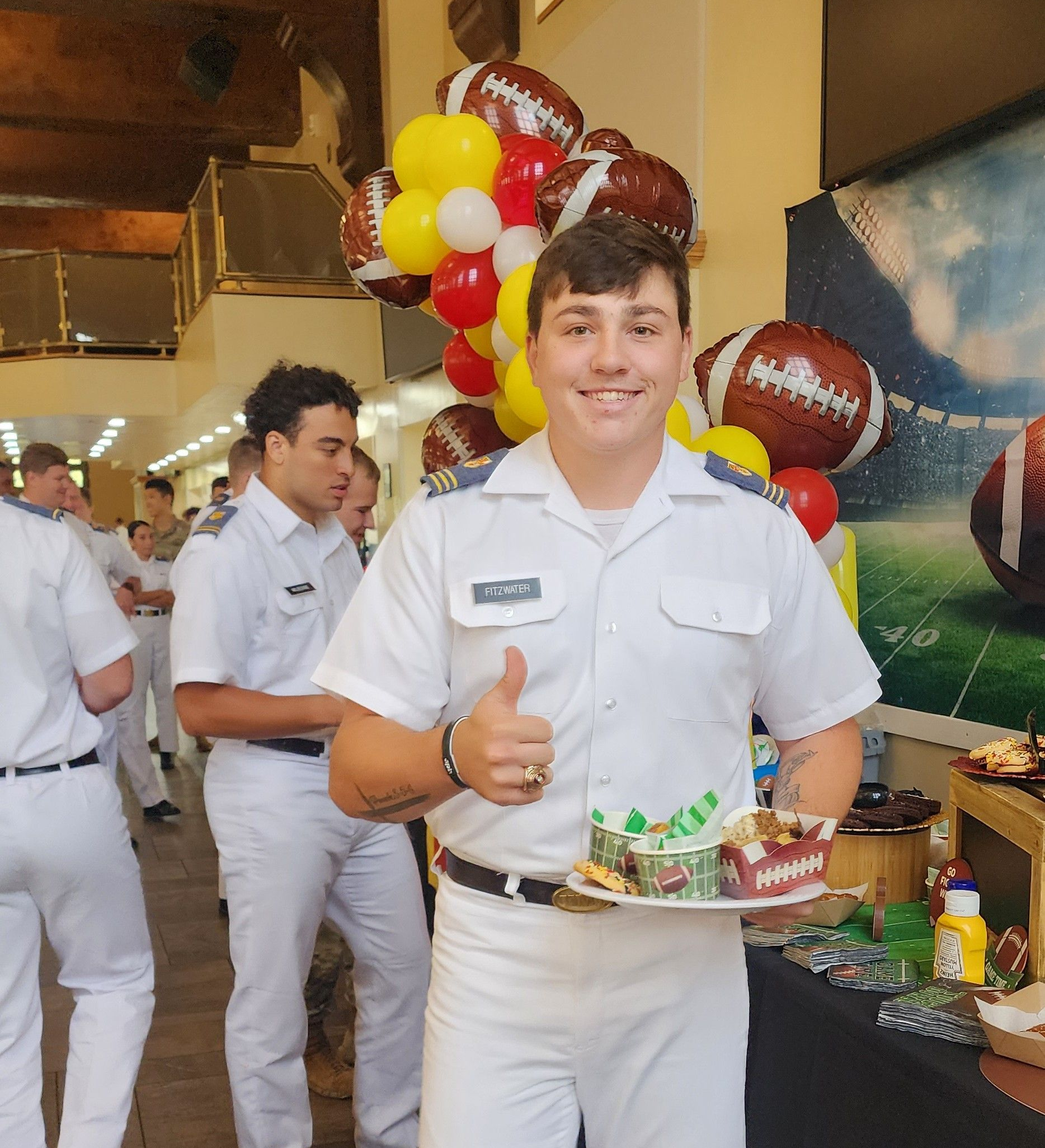 Man in uniform holding cake, giving thumbs up. Balloons and football decor in the background.