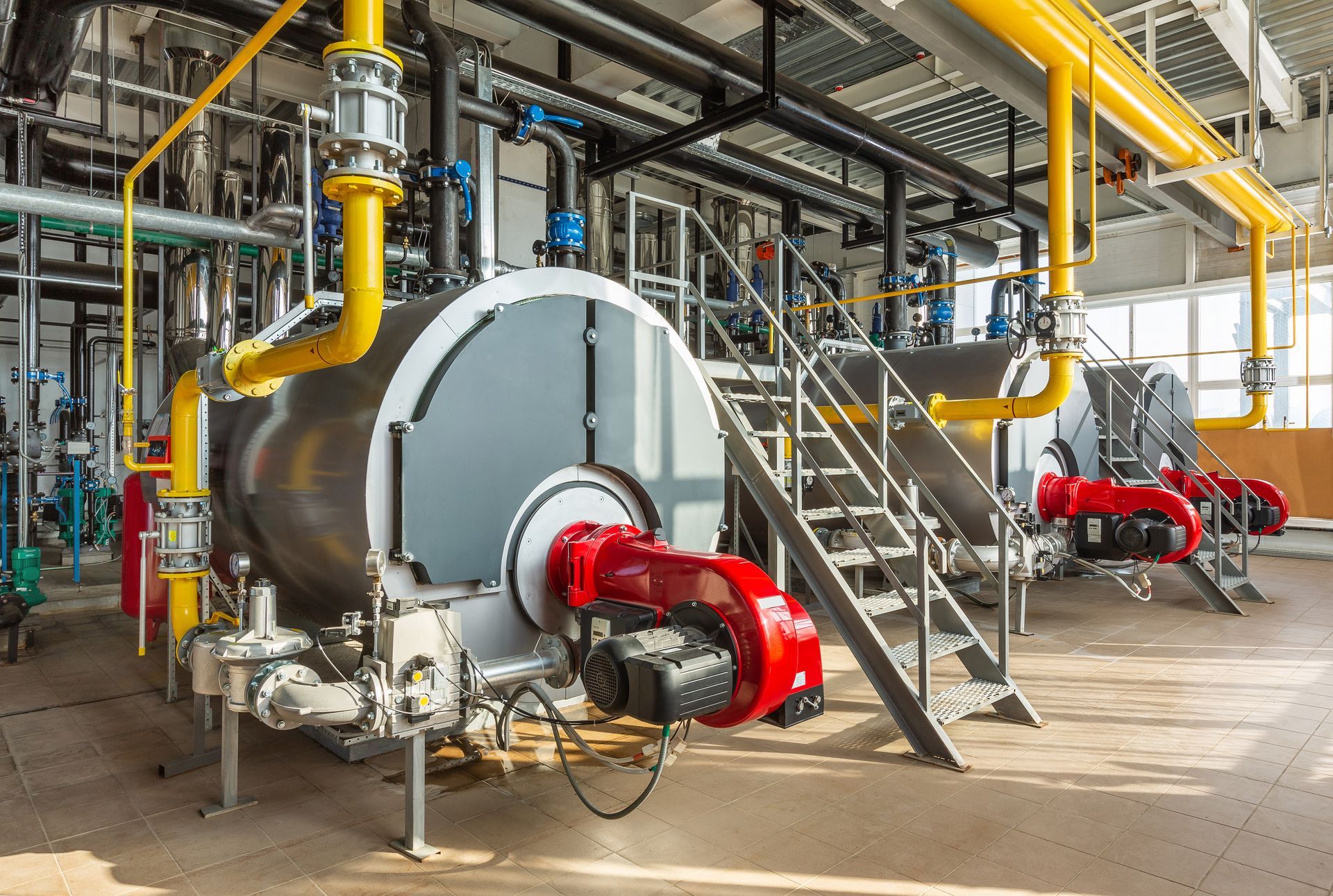 Large industrial boiler units with yellow pipes and red burners in a plant room.