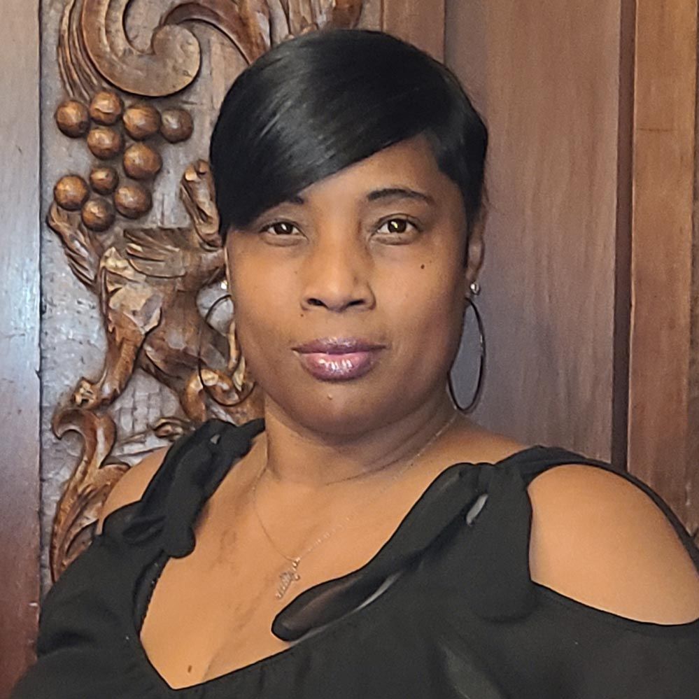 A woman in a black dress and hoop earrings is standing in front of a wooden wall.