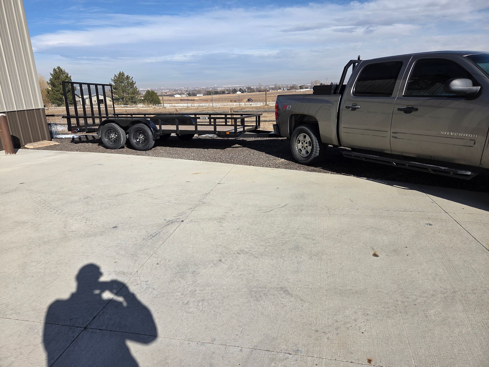 Pickup truck towing a flatbed trailer beside a building in a sunny gravel lot
