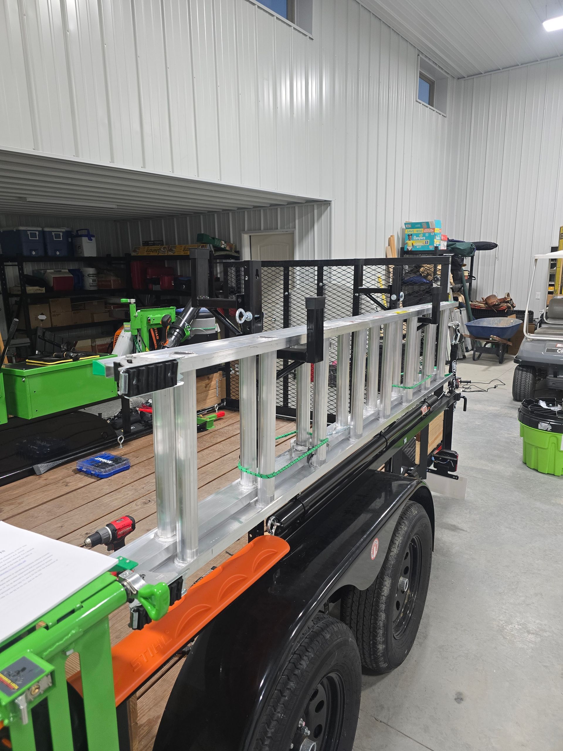 Aluminum ladder rack on a trailer in a workshop, with green and orange equipment visible.