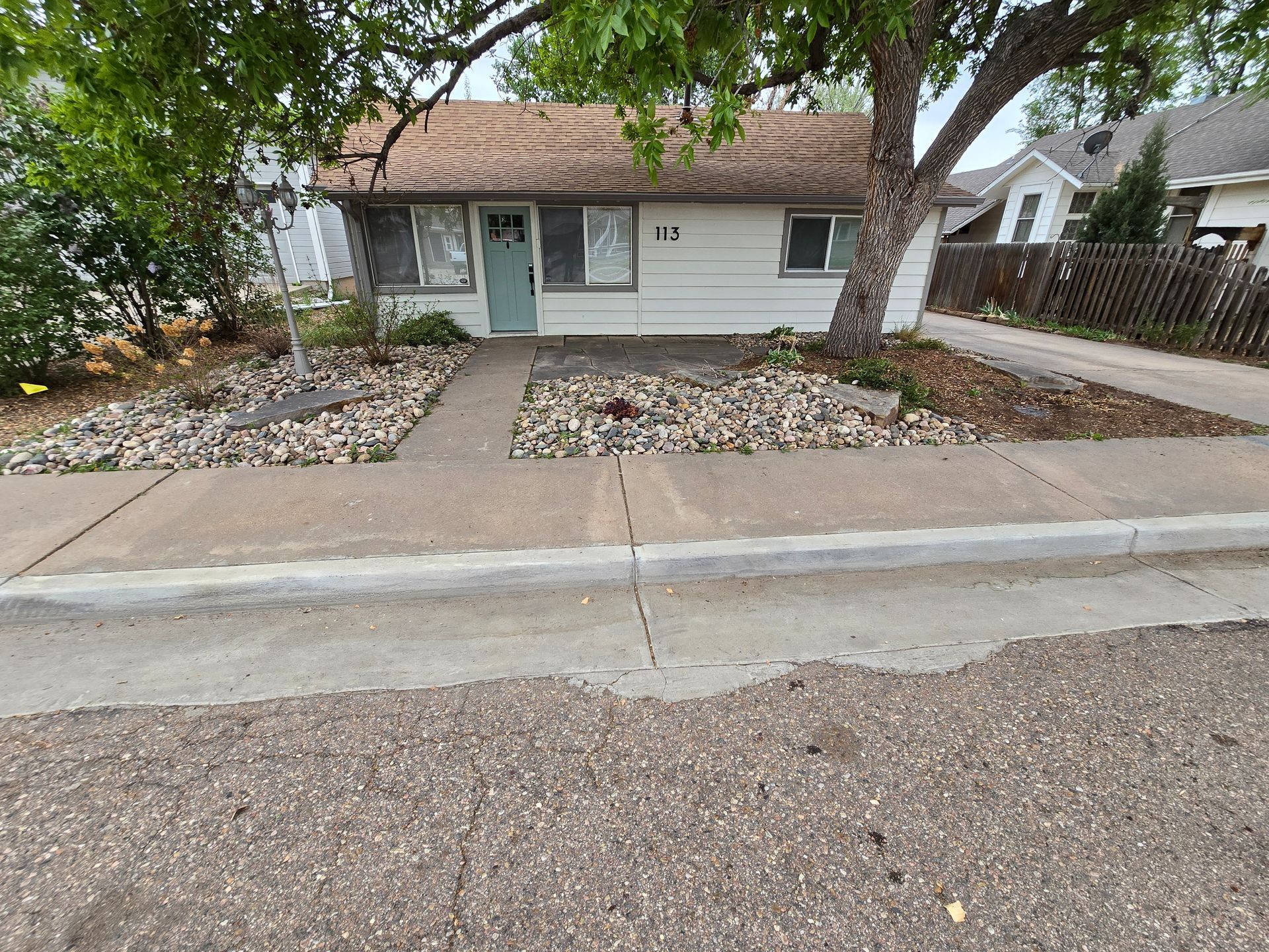 Single-story house with a front walkway, large trees, and rock landscaping by the street