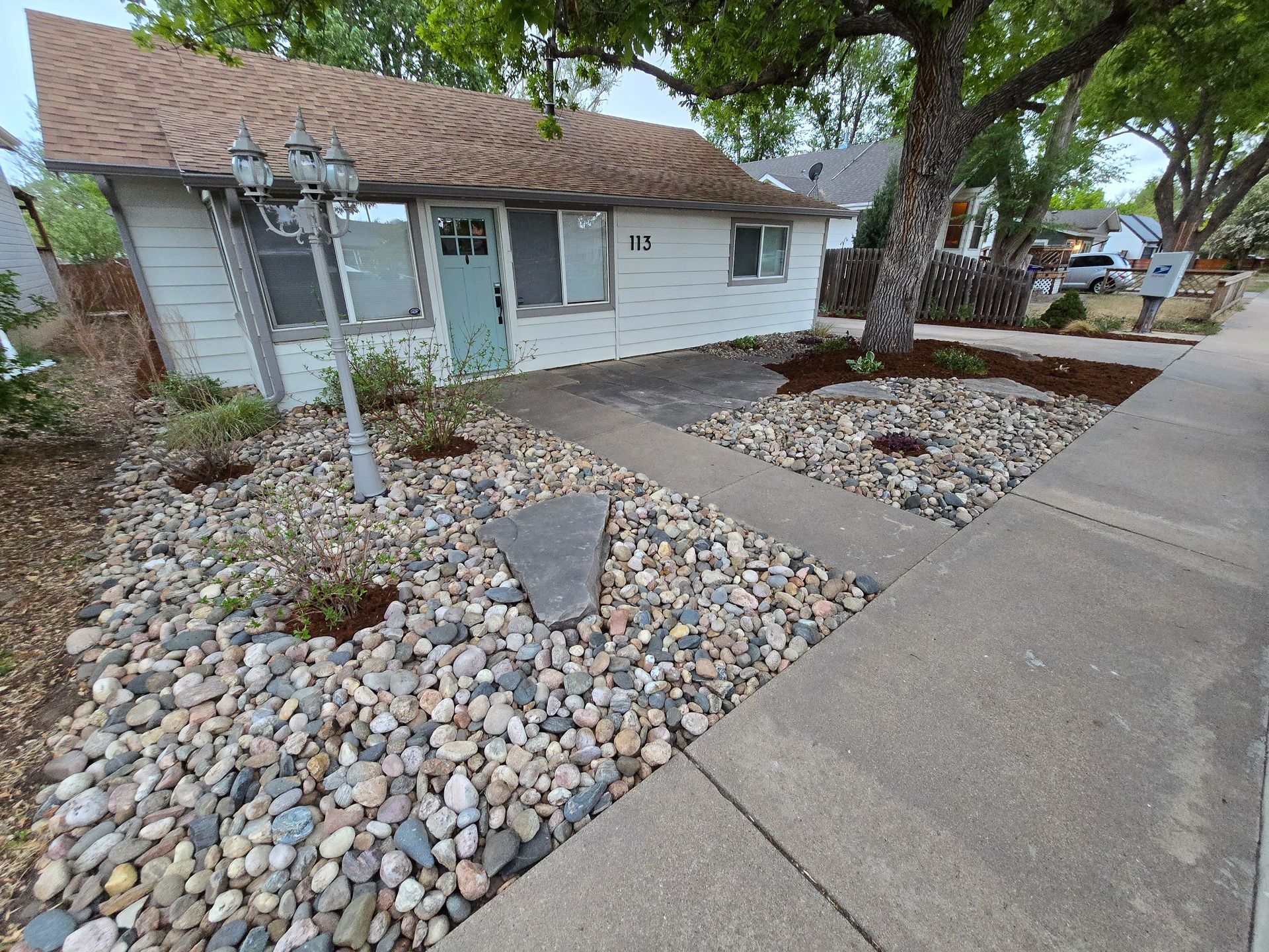 Front yard with rock landscaping, concrete walkway, large tree, and a single-story house with white siding.