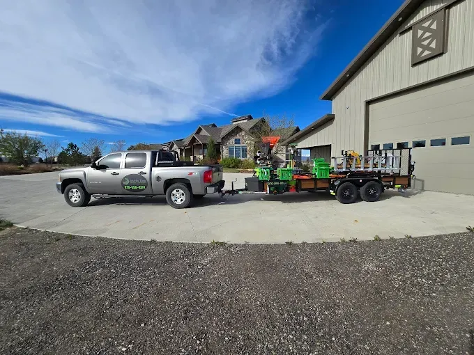 Silver pickup truck towing a green trailer beside a large beige barn under a bright blue sky