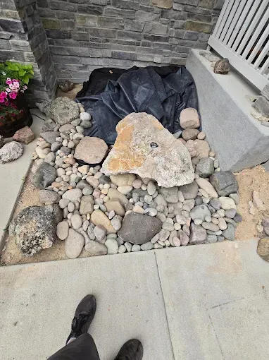 Rock garden bed with river stones, large flat rock, black tarp, beside a stone wall and concrete walkway