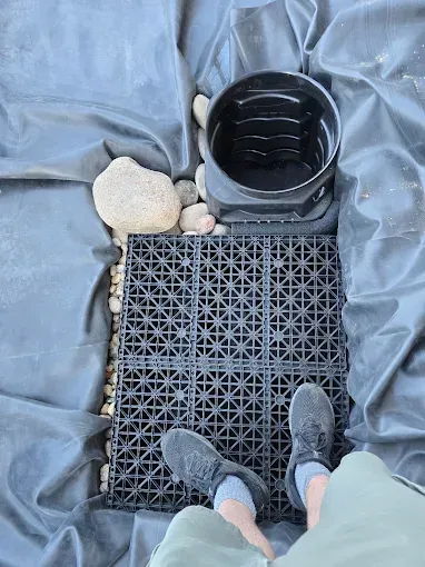 Top-down view of boots on a black grid mat beside a black bucket and white bagged material on gray tarp