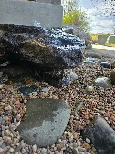 Decorative rock garden with dark boulders, pebbles, and a small puddle outdoors under a bright sky
