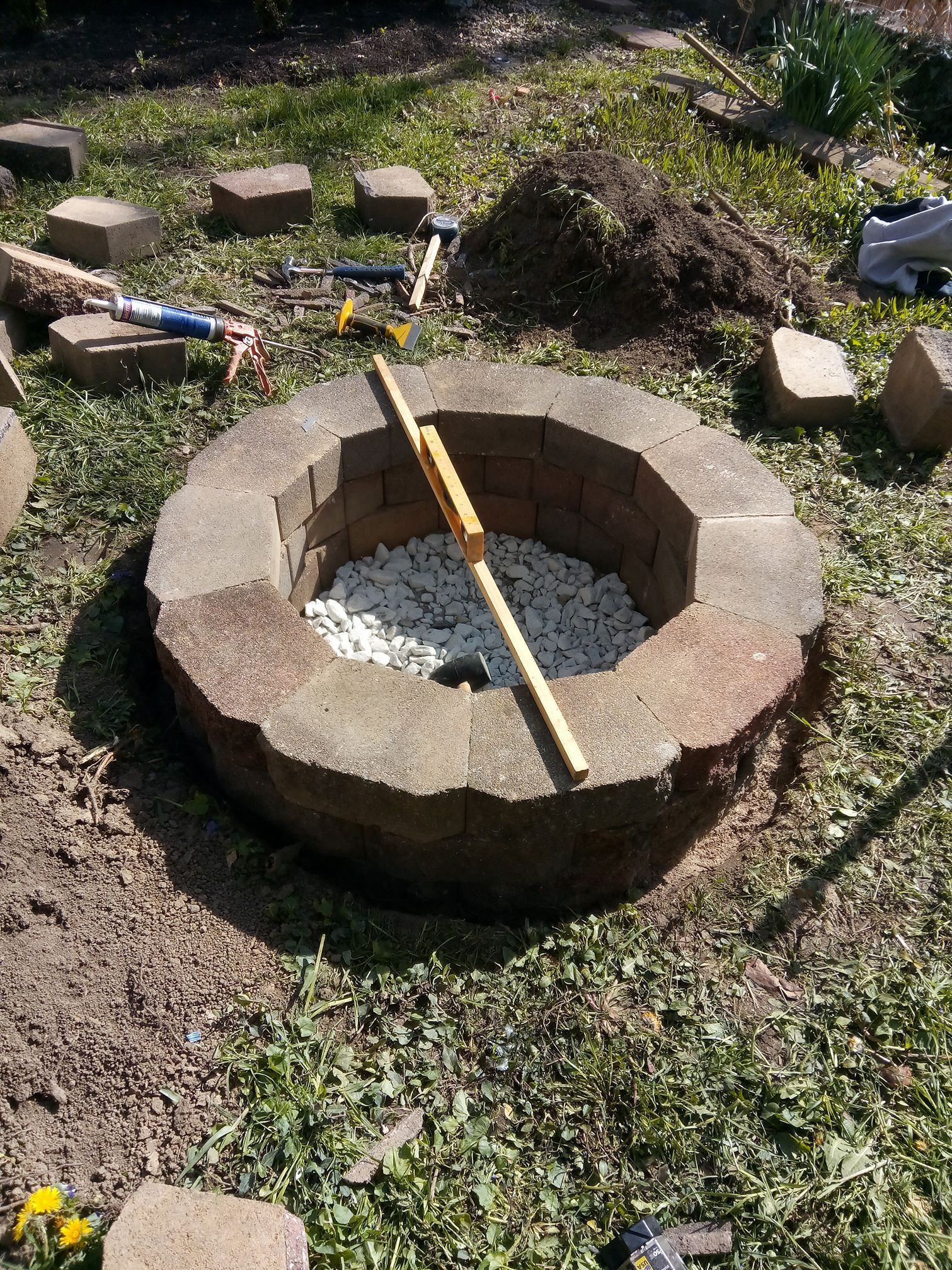 A circular stone fire pit under construction in a grass yard, featuring white gravel, loose pavers, and tools nearby.