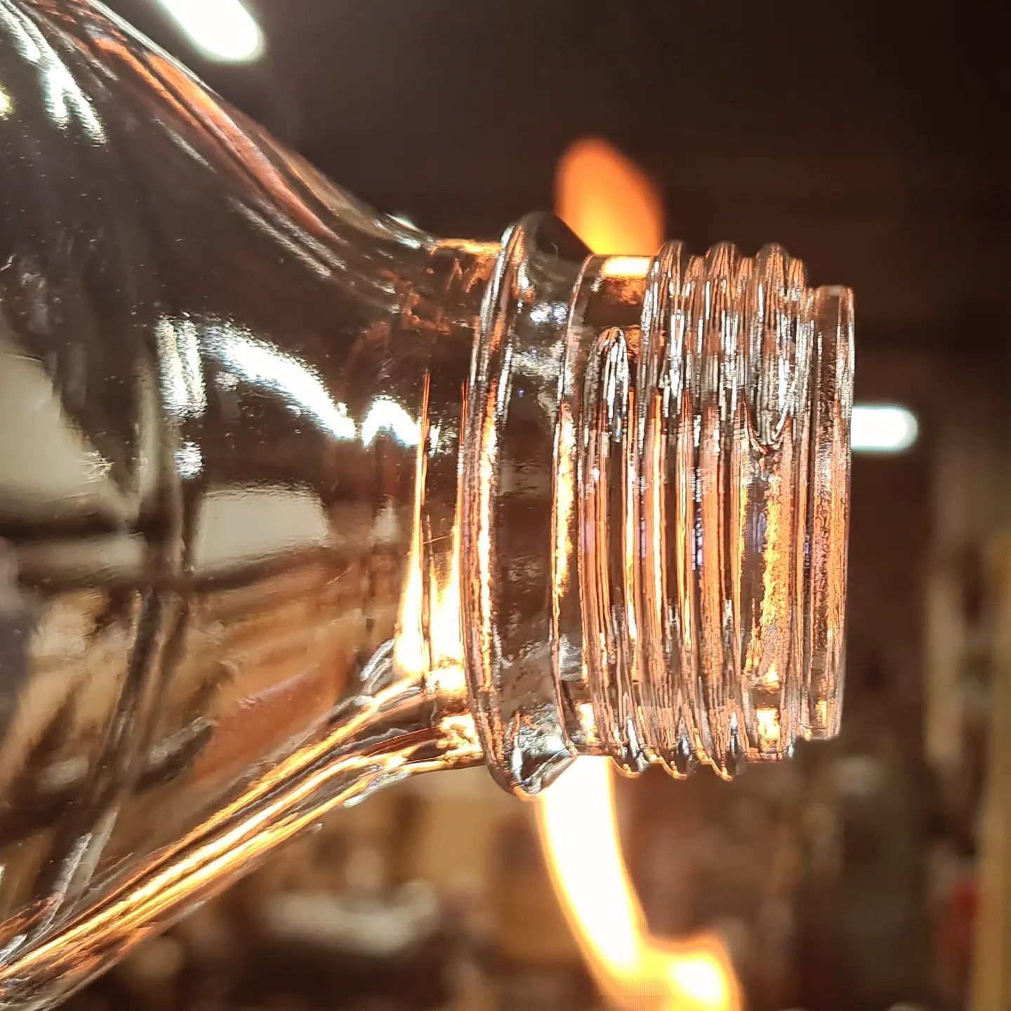 Primo piano del bordo di una bottiglia di vetro trasparente con fili visibili, illuminato da una fiamma arancione.