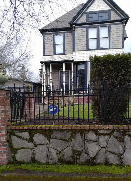 Victorian house with white siding and black trim behind a black iron fence and stone wall.