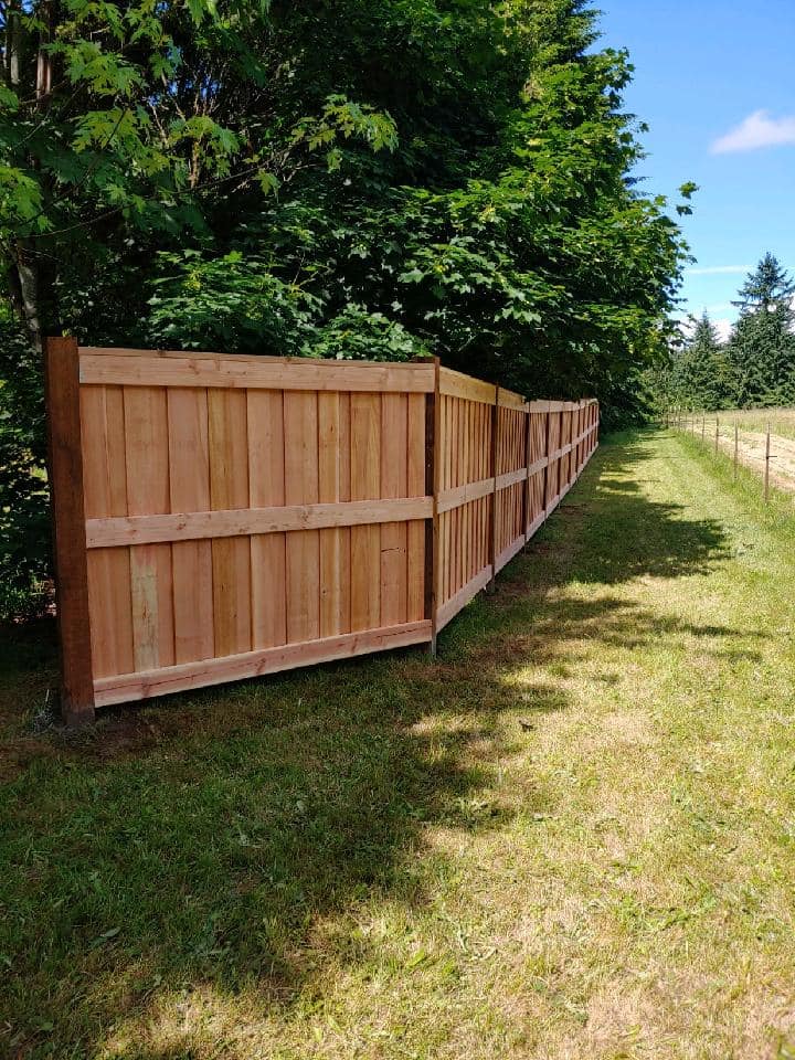 Wooden privacy fence beside a grassy path, with trees on one side and open field on the other.