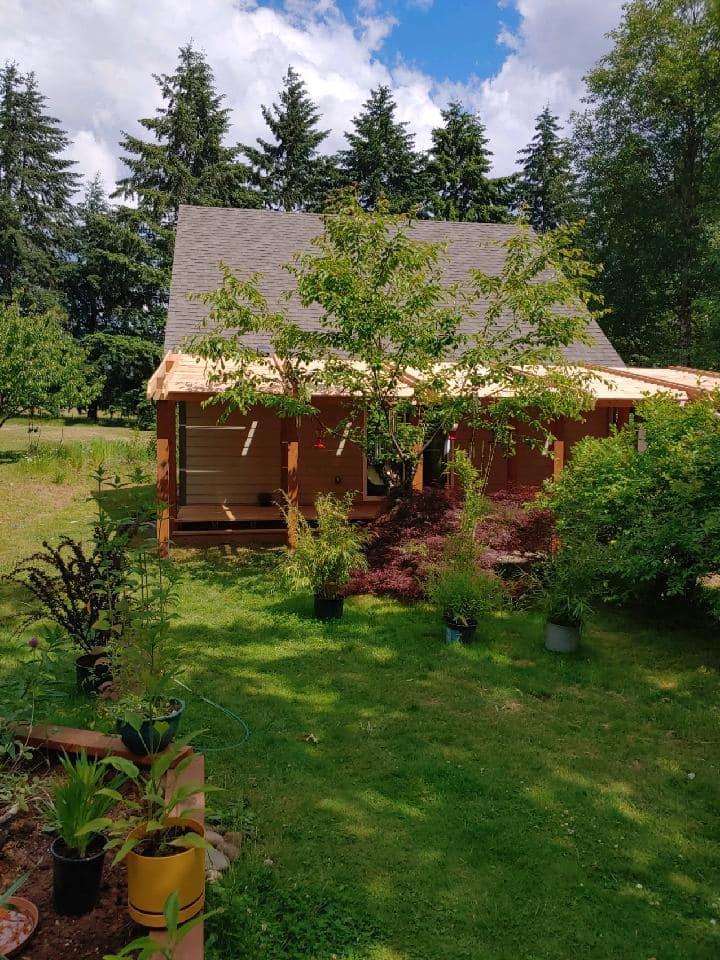Rustic cabin with a porch in a green yard, surrounded by trees under a bright sky.