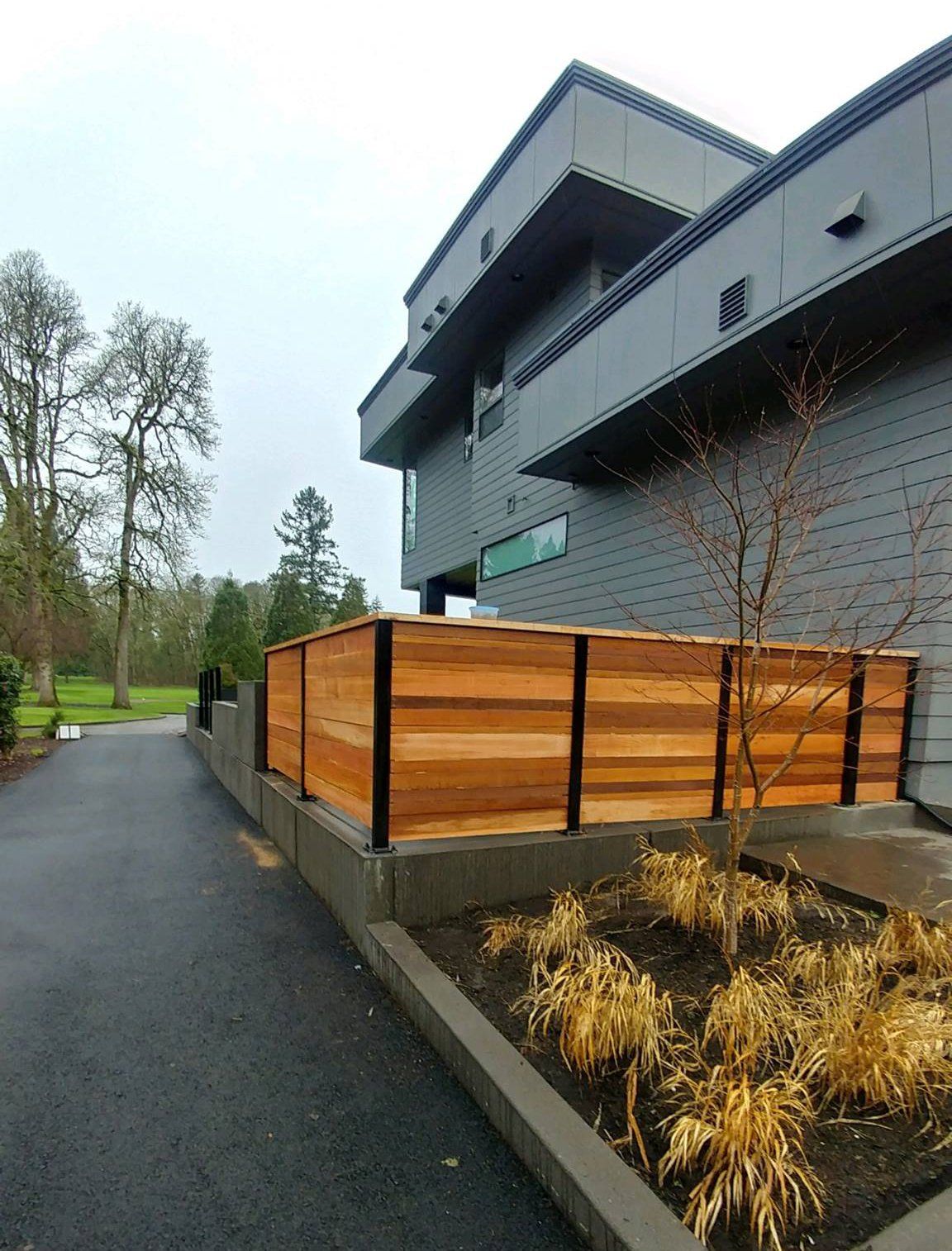 Modern gray house with a wooden fence beside a wet driveway and landscaped garden.