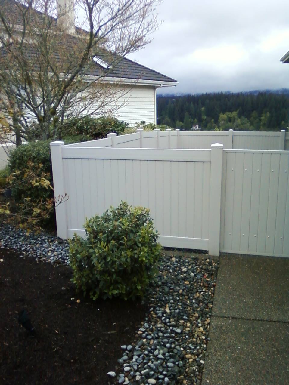 White vinyl fence beside a shrub and house, with gravel yard and cloudy hills in the background.