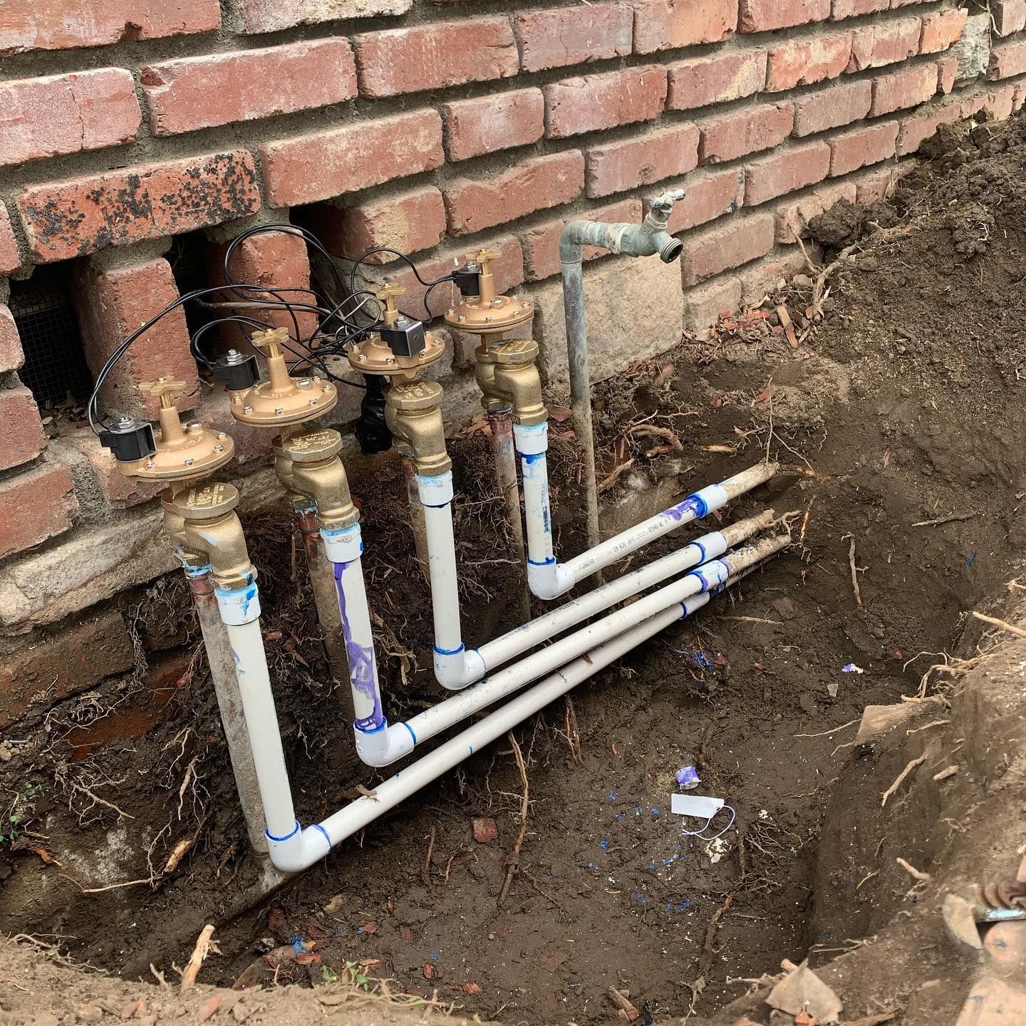 Irrigation system plumbing with PVC pipes and brass valves installed next to a brick foundation. 