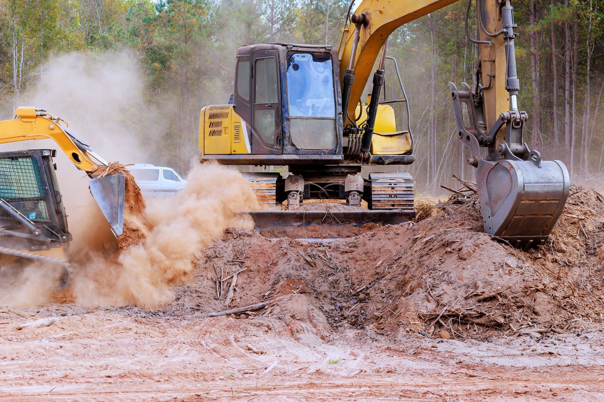 Two yellow excavators clear earth and brush in a wooded area, creating a large cloud of dust.