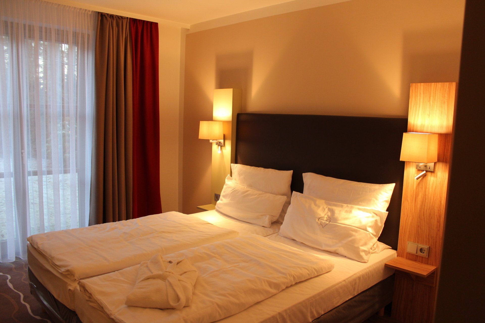 Modernes Hotelzimmer mit bezogenem Doppelbett, warmen Nachttischlampen und bodentiefem Fenster.