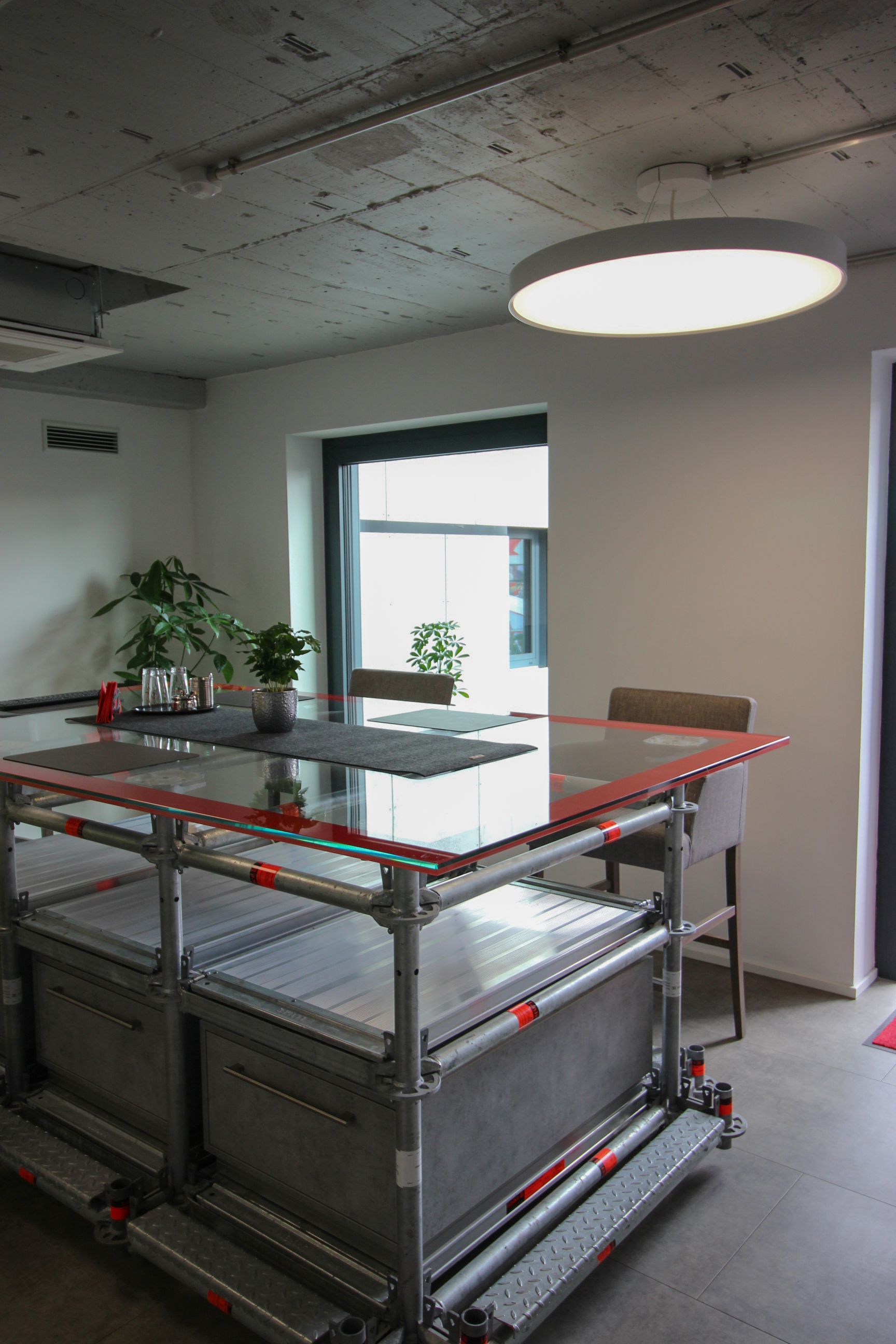 Industrielles Küchendesign: Eine großflächige, runde LED-Pendelleuchte leuchtet einen Glastisch von oben aus.