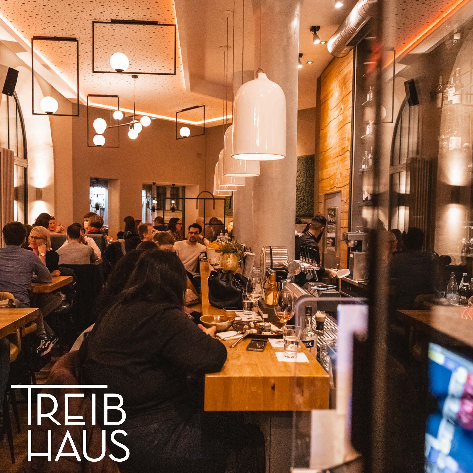 Belebte Bar-Beleuchtung: Große weiße Industrie-Hängeleuchten und schwarze Rahmenleuchten erhellen den langen Tresen.