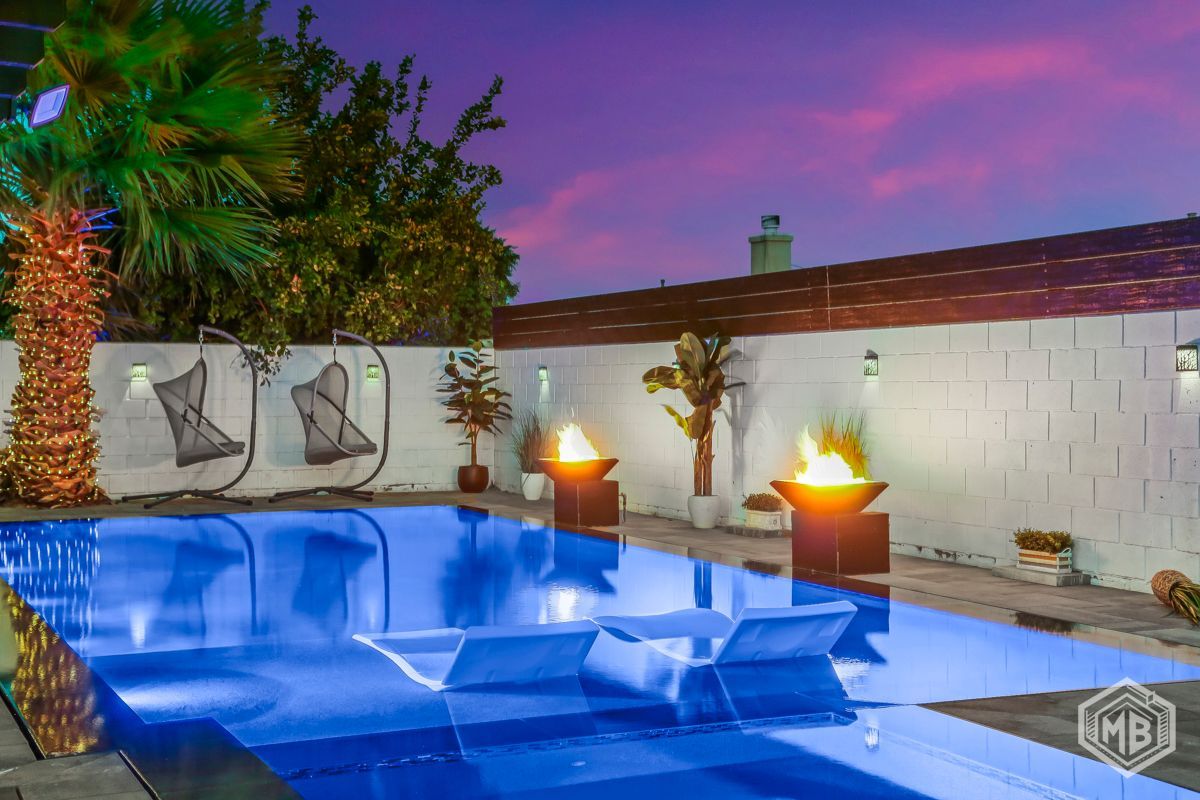 A swimming pool at sunset, featuring two fire pits, lounge chairs in the water, and hanging chairs against a white wall.