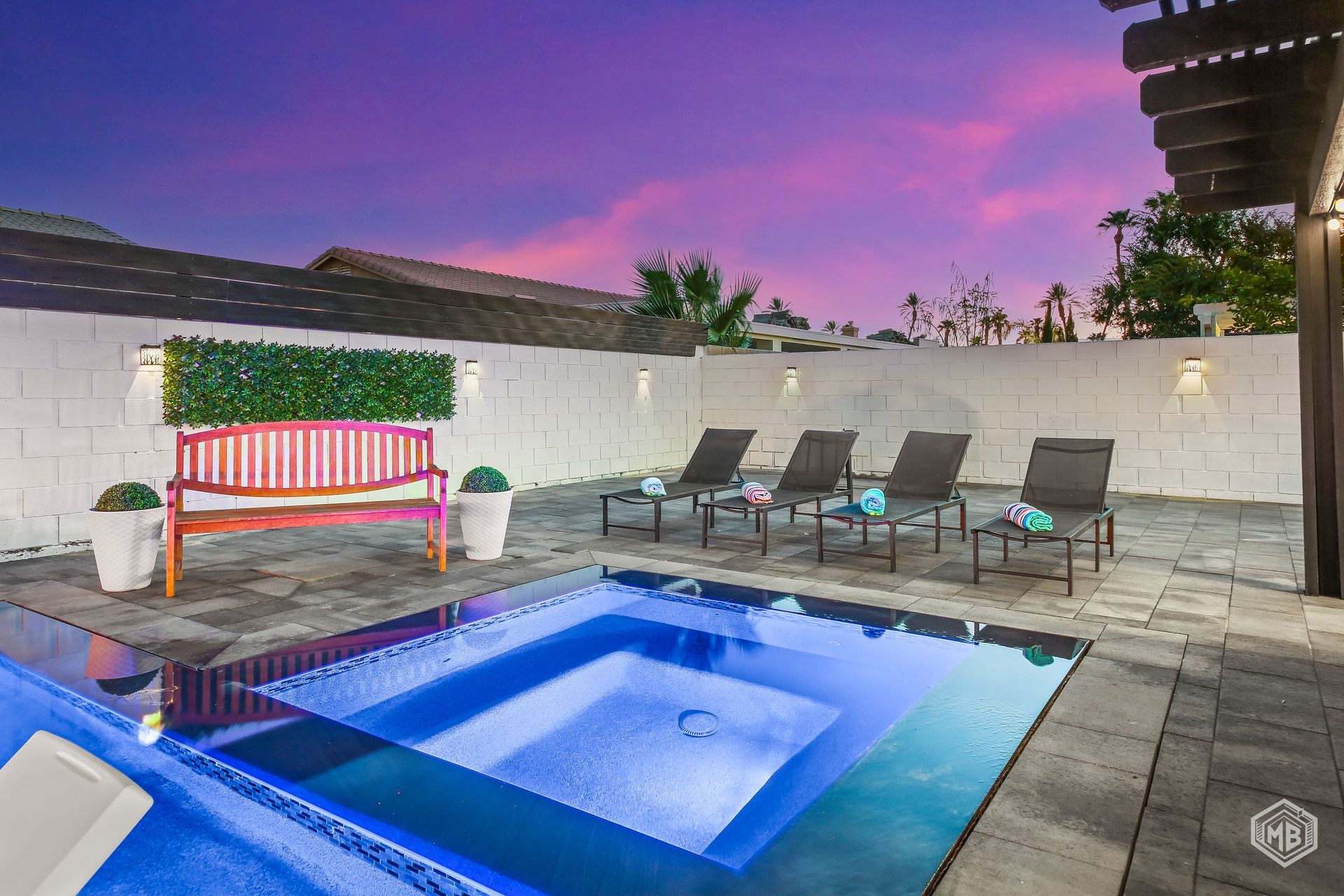 A backyard swimming pool at sunset, featuring a pink bench, loungers, and a white privacy wall against a purple sky.