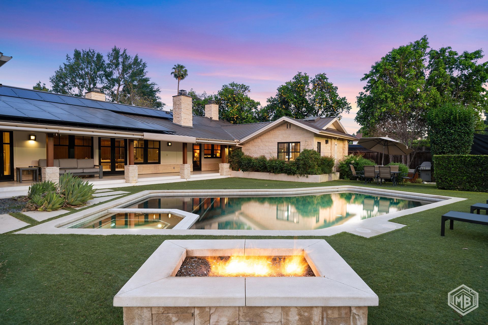 A backyard with a stone fire pit in the foreground, a swimming pool, and a single-story house at sunset.