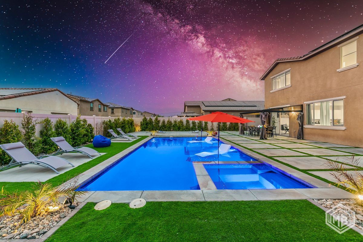 A modern backyard pool and patio at night, with lounge chairs and a bright red umbrella under a vibrant starry sky.