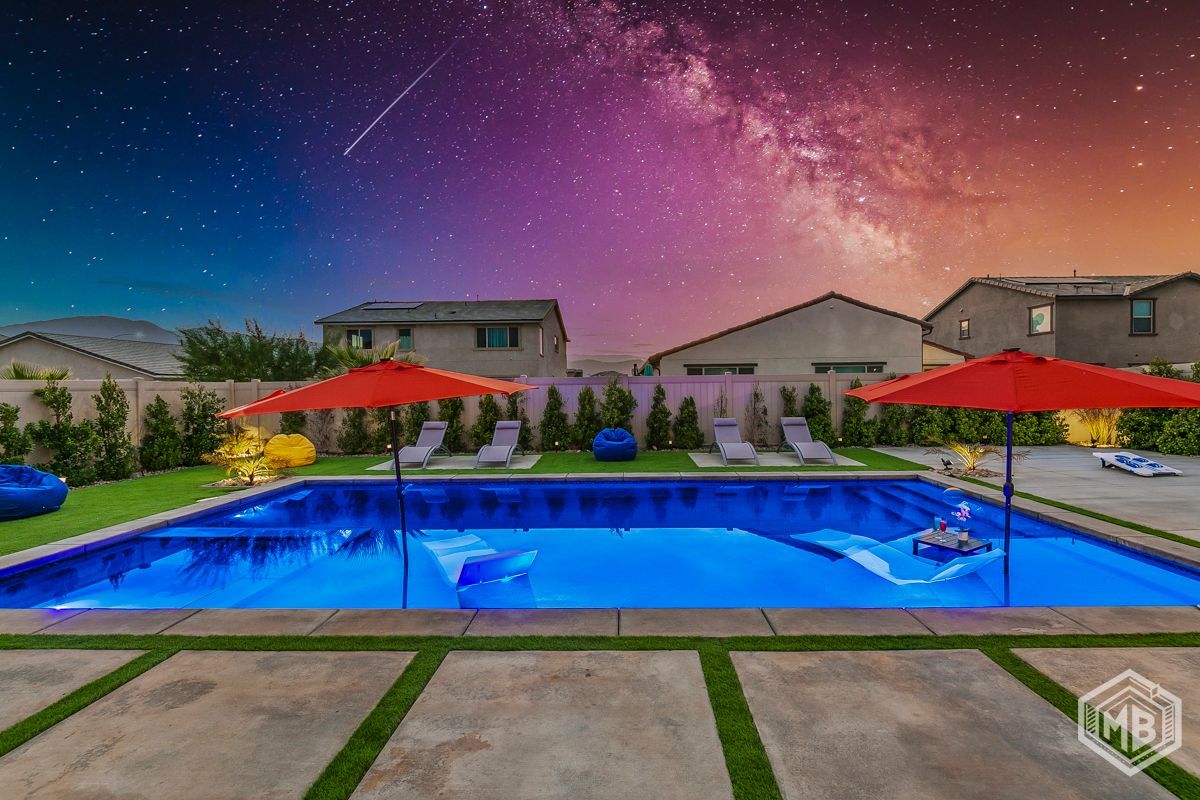 A swimming pool in a backyard at night, illuminated by blue lights under a vibrant, star-filled sky.