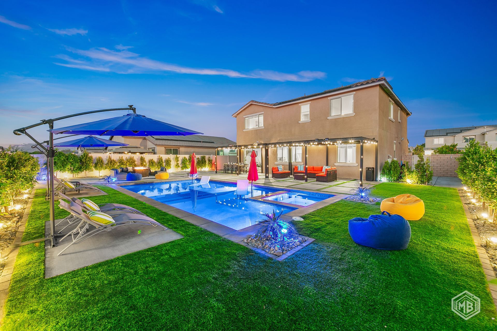 A backyard at dusk featuring a lighted swimming pool, patio furniture, a blue umbrella, and beanbag chairs on green grass.