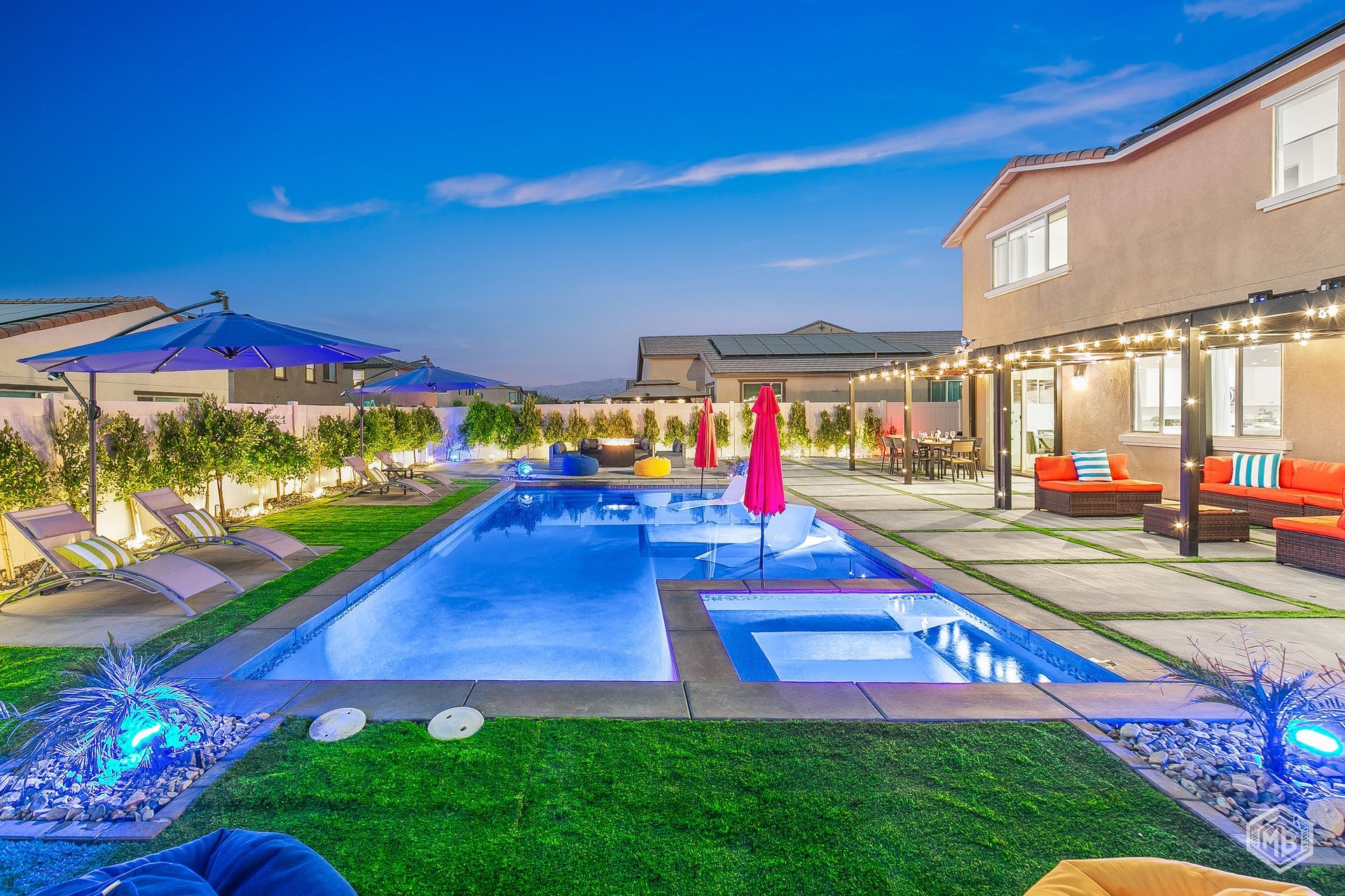 A modern backyard at dusk featuring an illuminated swimming pool, a hot tub, patio furniture, and string lights.