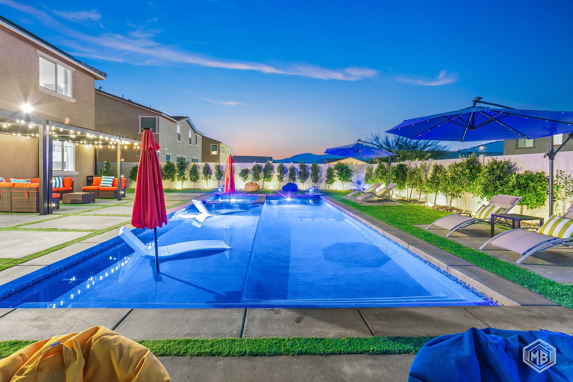 A backyard swimming pool illuminated at twilight, featuring a red umbrella, lounge chairs, and string lights on a patio.
