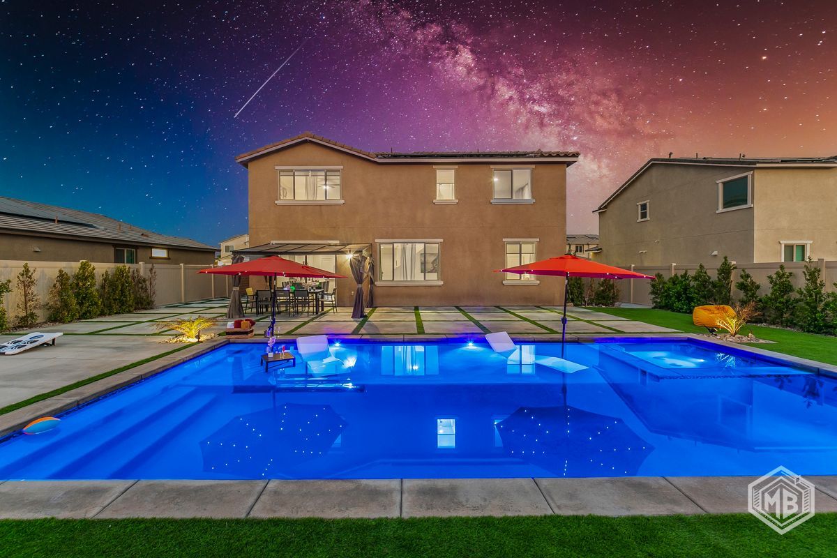 A modern two-story suburban house at night with a glowing blue backyard swimming pool under a vibrant starry sky.