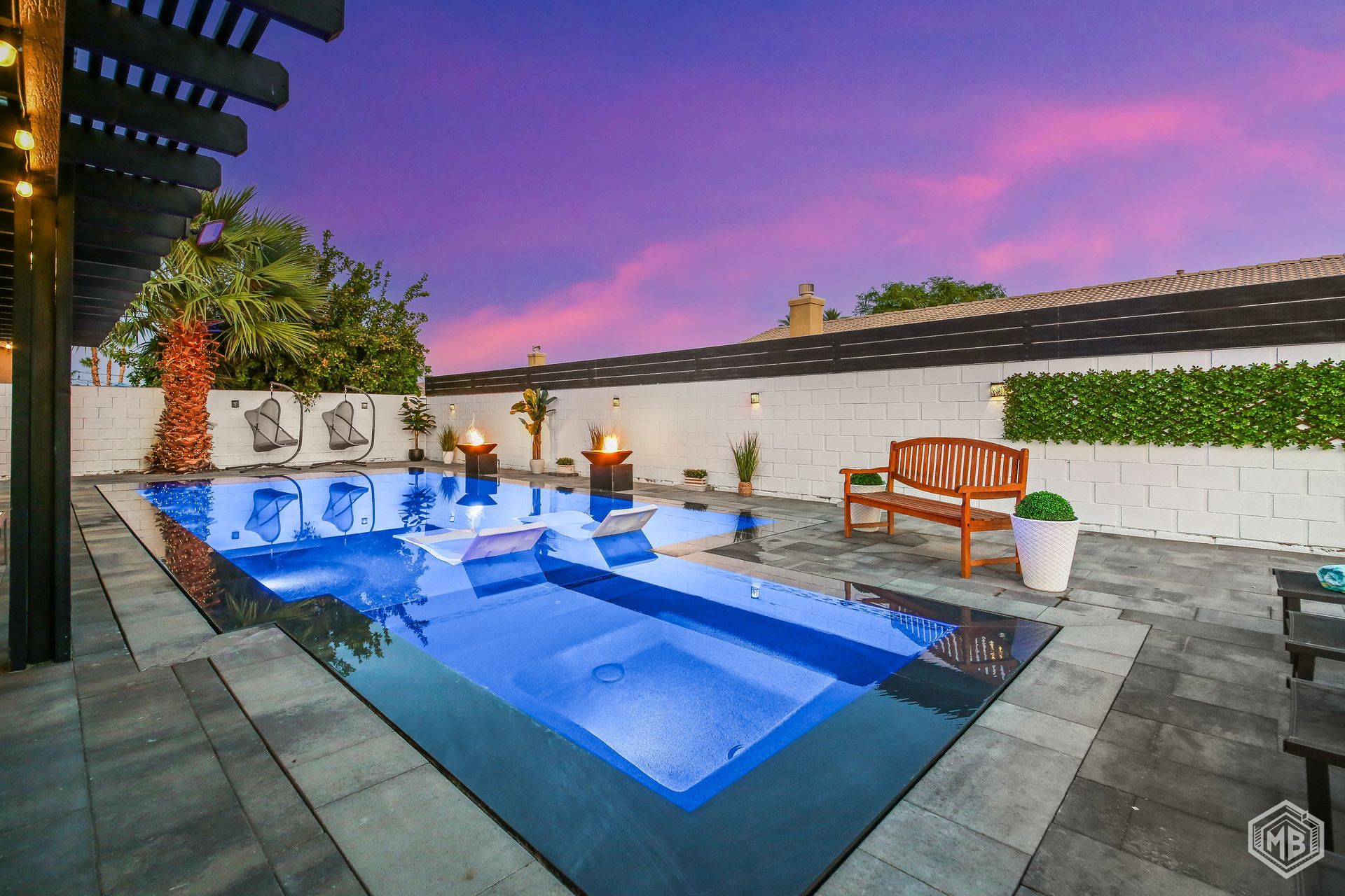 A rectangular backyard swimming pool with dark tile surround, a wooden bench, and a sunset sky overhead.
