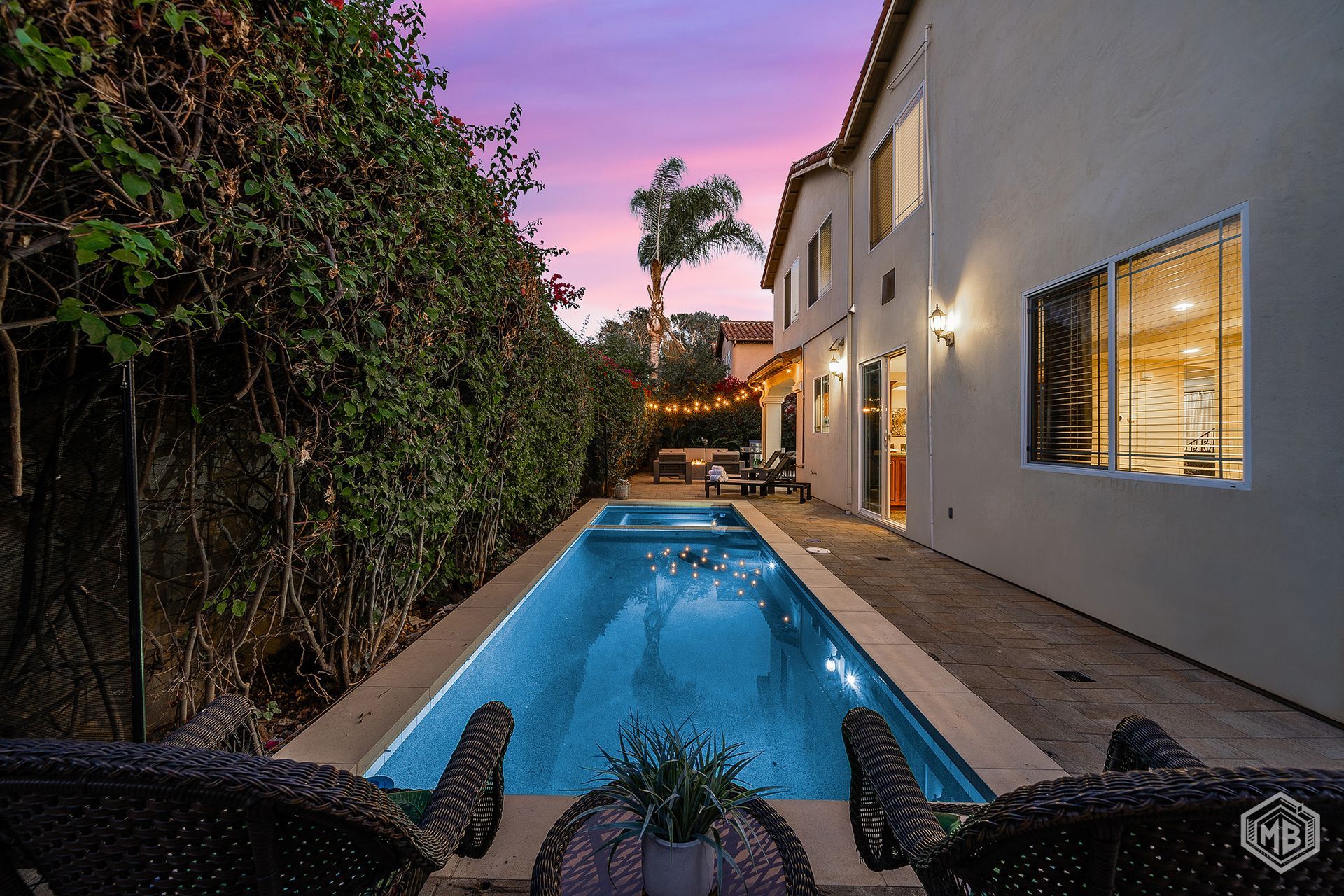 A narrow rectangular swimming pool illuminated by string lights at dusk, set between a house and a dense hedge.