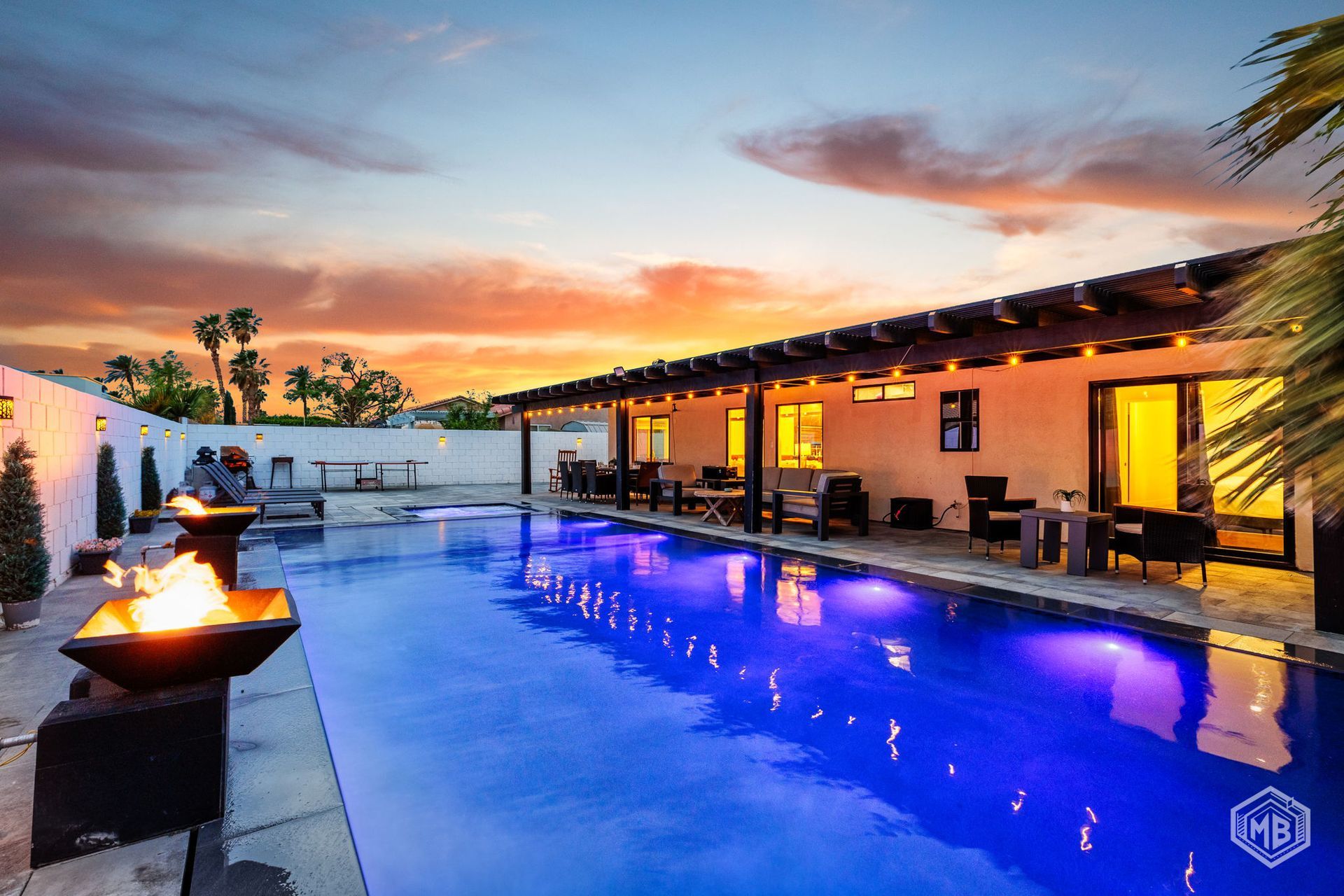 An outdoor swimming pool at sunset, featuring a glowing fire pit and a poolside patio with lighting under a wood pergola.