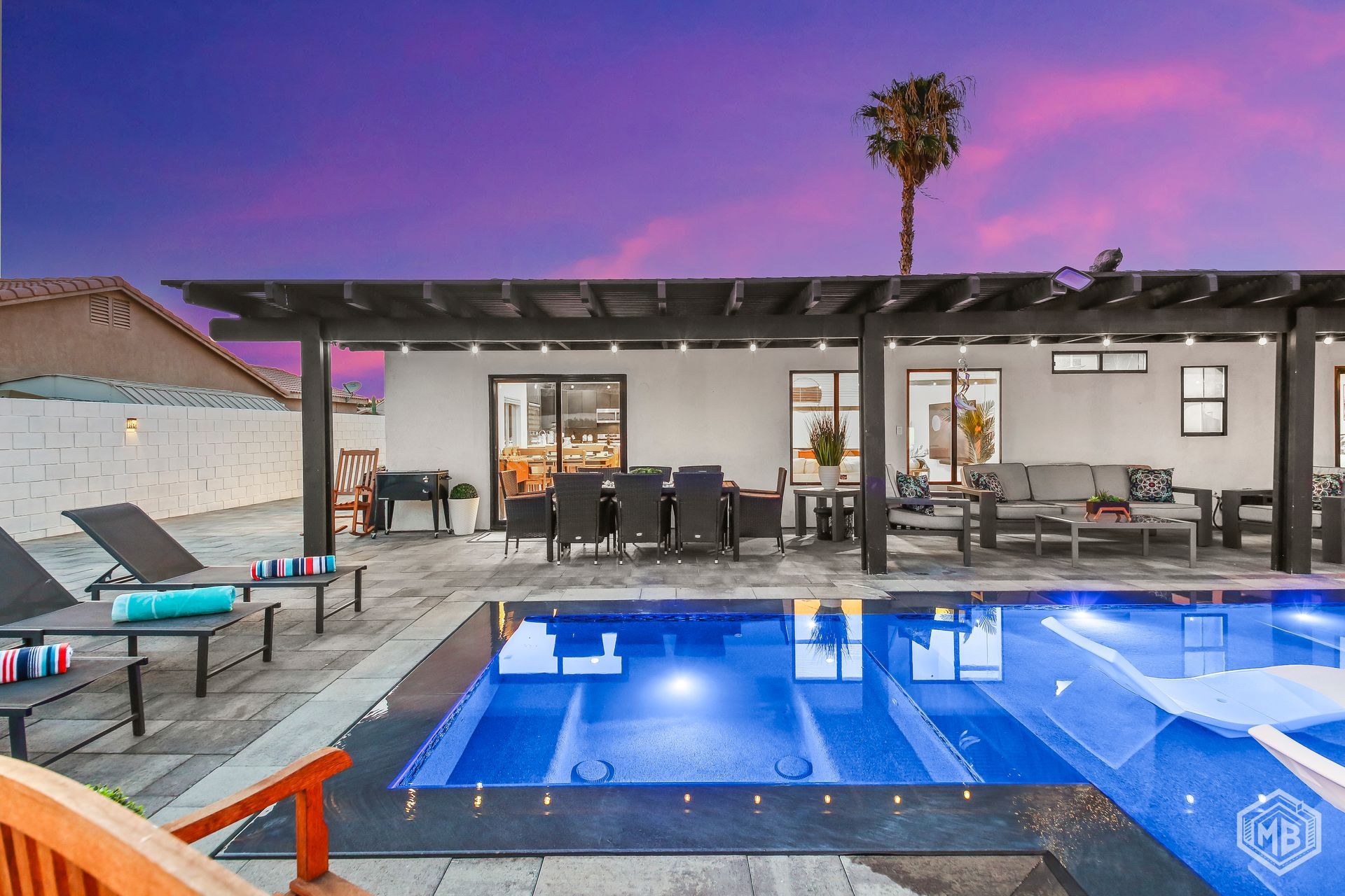 A backyard patio at sunset with a pool, outdoor dining table, lounge chairs, and a covered pergola.