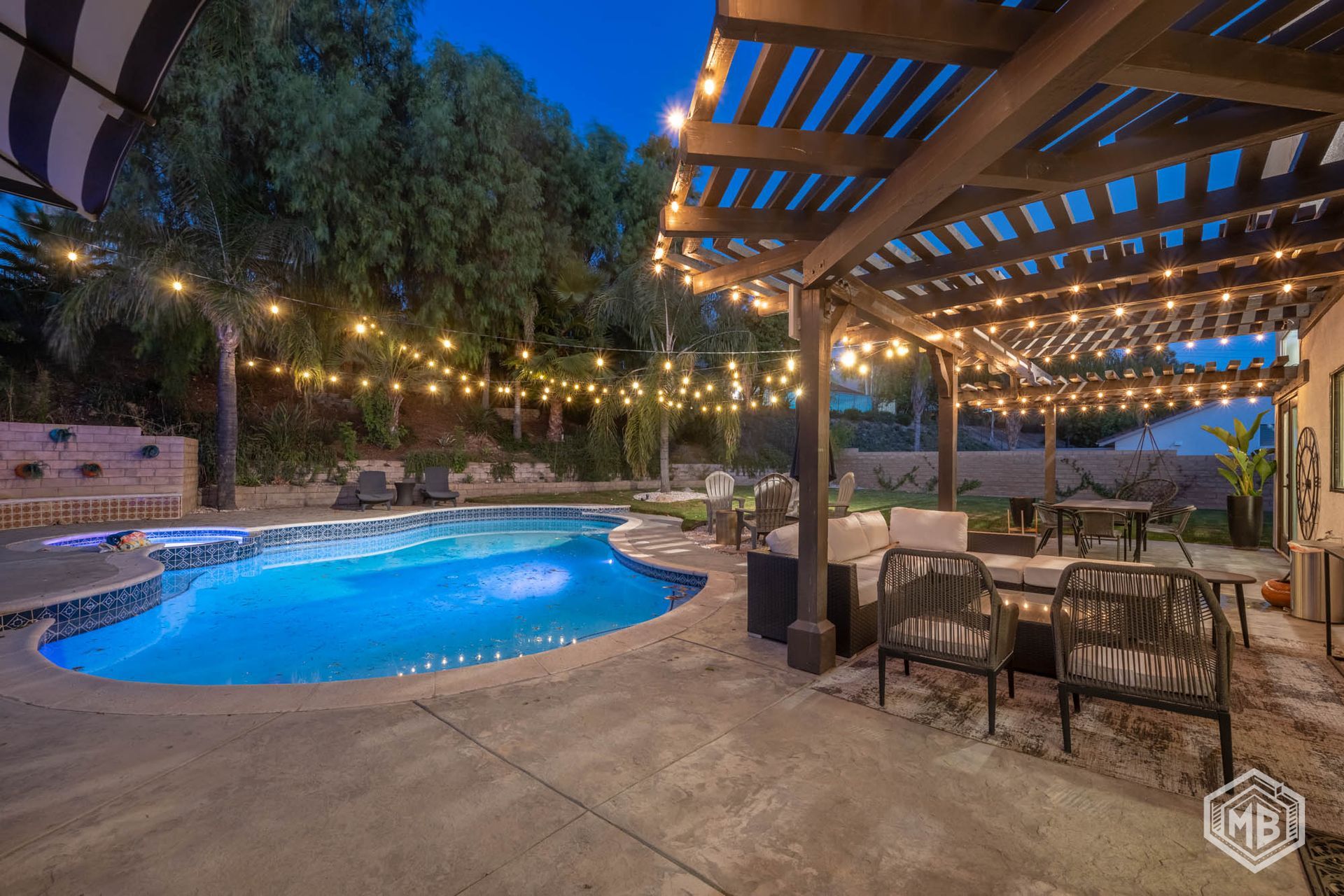 A twilight view of a swimming pool with string lights draped over a patio area featuring outdoor furniture and a pergola.