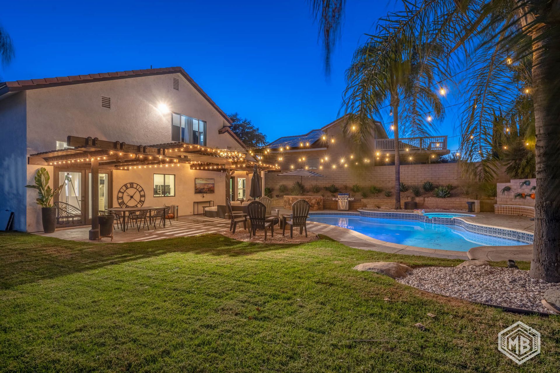 A twilight view of a house backyard featuring a swimming pool, a covered patio with dining furniture, and string lights.
