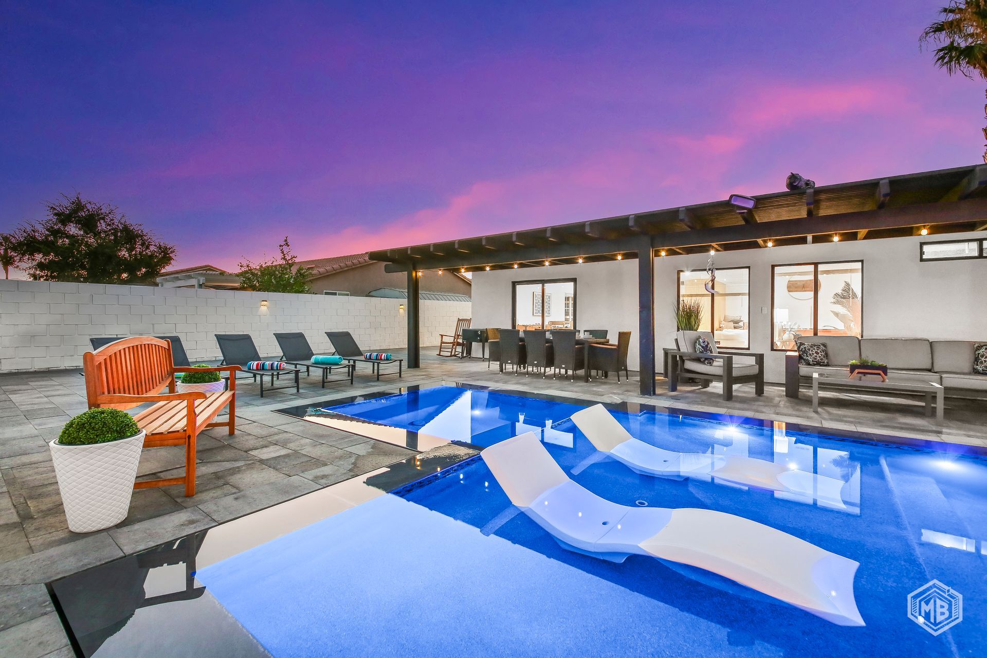 A backyard pool with submerged lounge chairs under a pergola, set against a vibrant sunset sky.