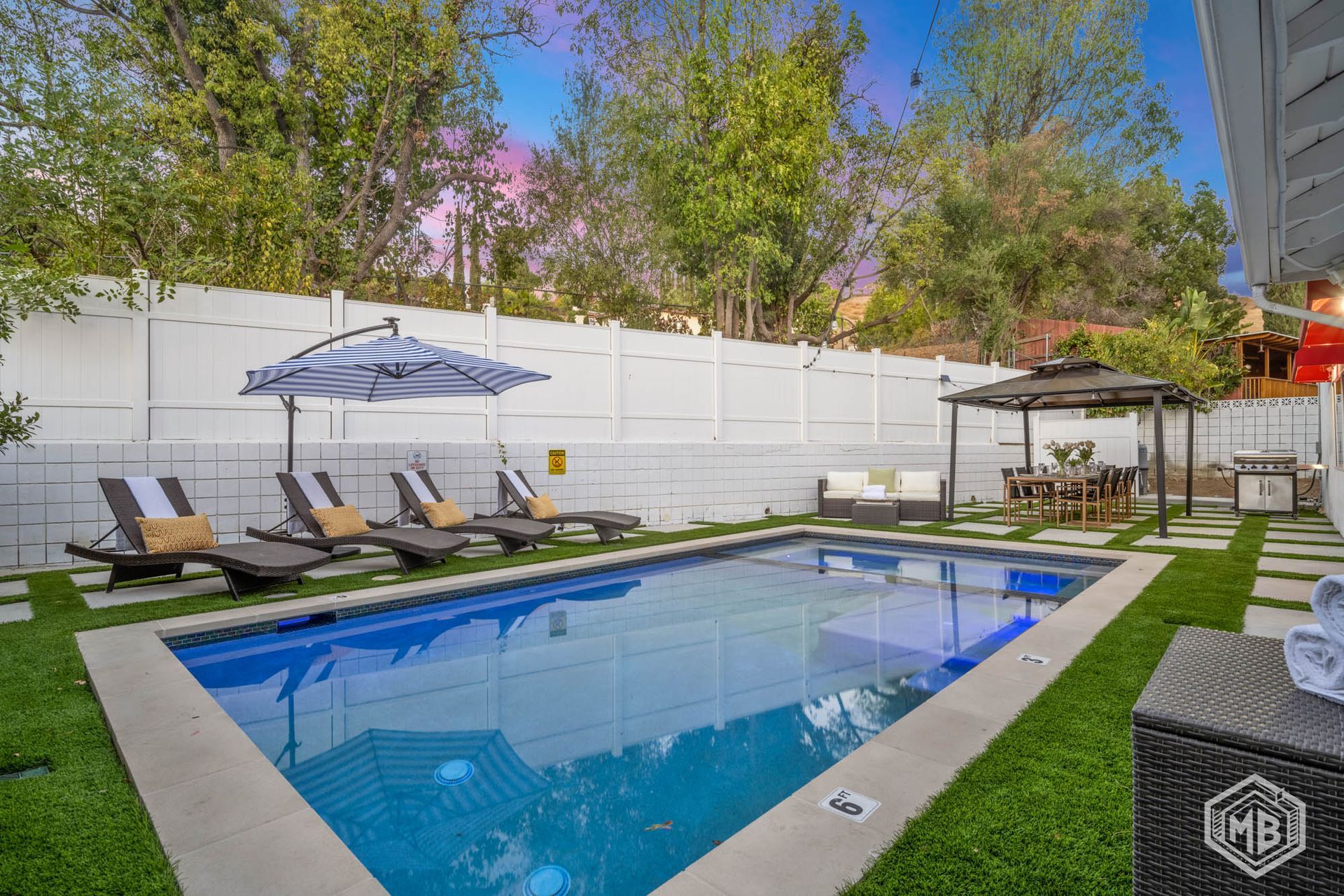 A rectangular backyard swimming pool with lounge chairs, a patio umbrella, and a gazebo on artificial grass.