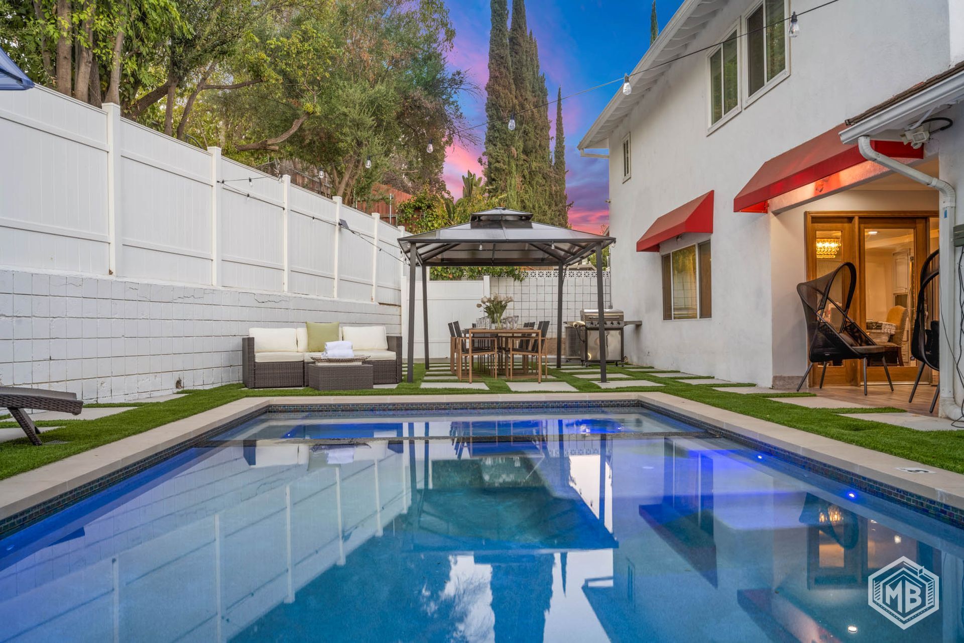 A backyard swimming pool with a gazebo, outdoor seating, and a house exterior at twilight.